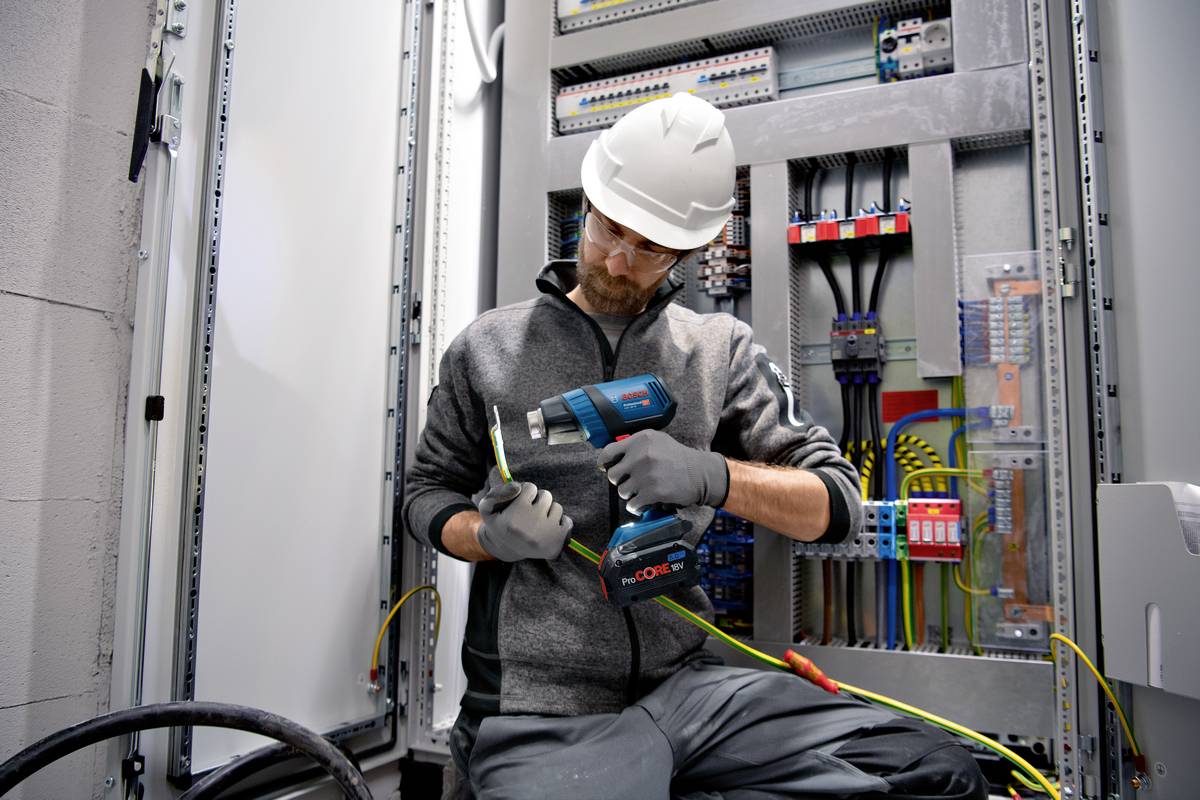 Ein Elektriker in Schutzausrüstung arbeitet mit einem Elektrowerkzeug an einem offenen Sicherungskasten, konzentriert bei der Arbeit.