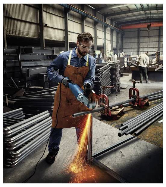 Ein Arbeiter in einer Werkstatt benutzt ein Elektrowerkzeug, um Metall zu schneiden, wobei Funken durch die Luft sprühen. Im Hintergrund sind Stapel von Metallrohren zu sehen.