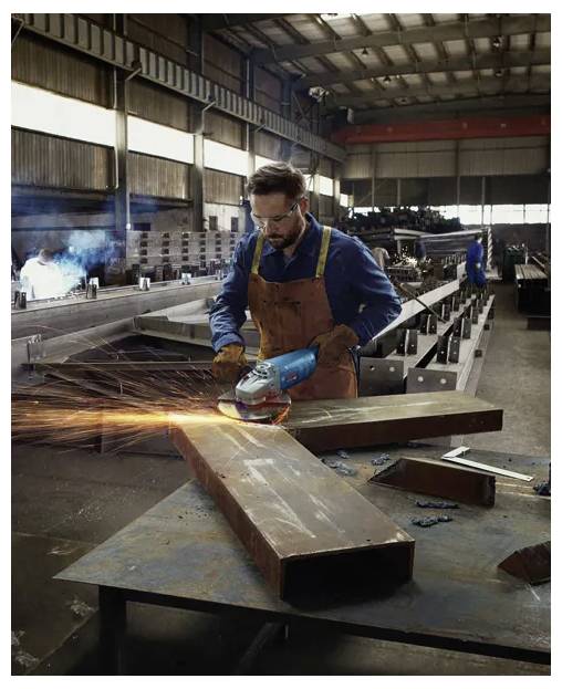 Ein Arbeiter in einer Werkstatt, ausgerüstet mit Sicherheitsausrüstung, schleift einen großen Metallträger und erzeugt dabei helle Funken. Im Hintergrund sind Metallstrukturen und Maschinen zu sehen.