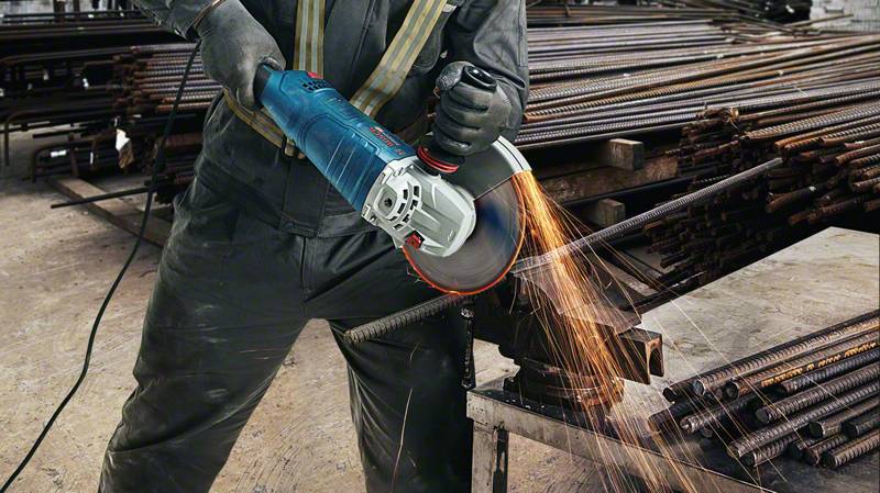 Arbeiter schneidet Metallstange mit Winkelschleifer, Funken fliegen. Im Hintergrund liegen weitere Metallstäbe aufgestapelt.