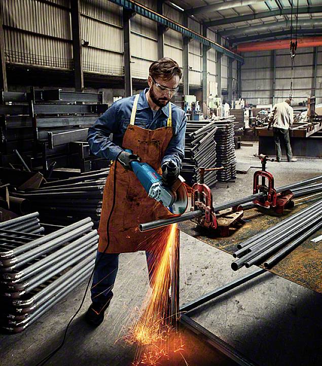 Ein Mann in einer Werkstatt schneidet Metallrohre mit einem Winkelschleifer. Funken fliegen, während er arbeitet. Im Hintergrund stehen mehr Rohre.