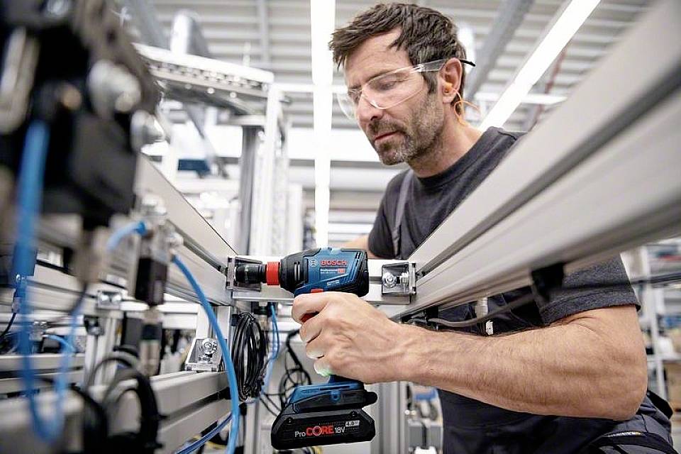 Ein Mann mit Schutzbrille arbeitet konzentriert an einer Maschine in einer Fabrik und benutzt dabei einen Akkuschrauber.