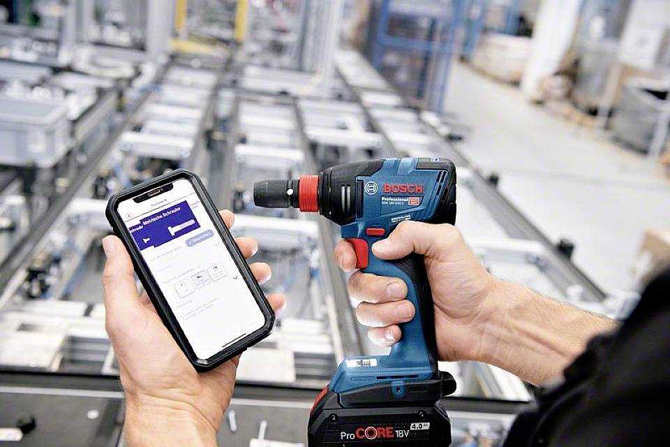 Ein Mensch hält einen Bosch-Akkubohrschrauber in einer Hand und ein Smartphone mit einer App in der anderen Hand in einer Werkstattumgebung.