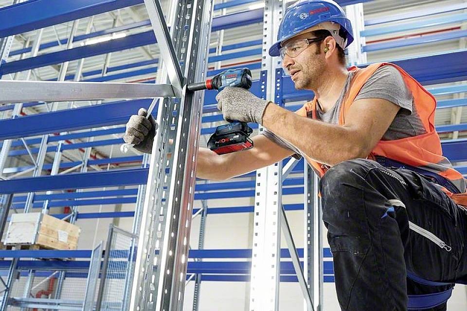 Ein Bauarbeiter in Sicherheitskleidung verwendet eine elektrische Bohrmaschine, um Metallgerüste in einem Lagerhaus zusammenzubauen.