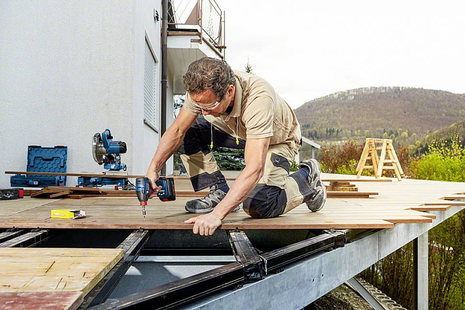 Ein Handwerker kniet auf einem Holzdeck und verwendet einen Akkuschrauber, um Platten zu befestigen. Werkzeugkoffer und Natur im Hintergrund.