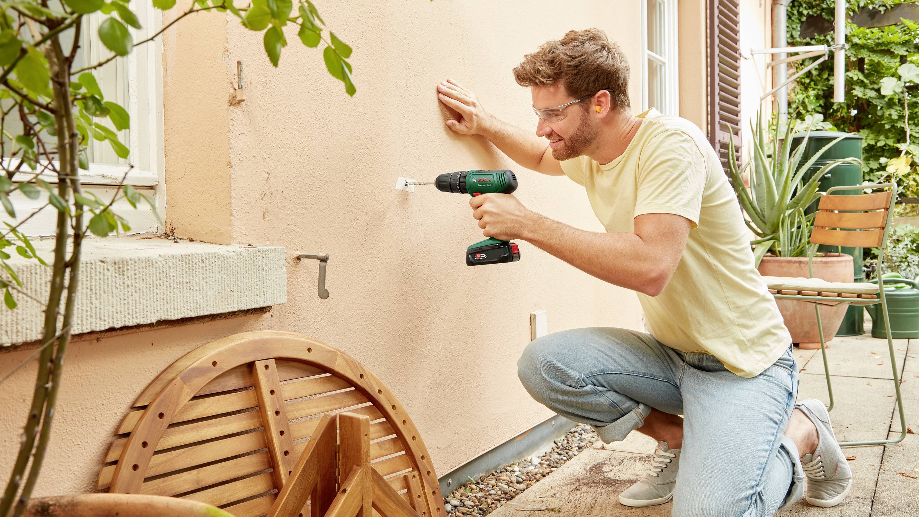 Ein Mann kniet draußen und bohrt ein Loch in eine Hauswand. Neben ihm stehen Pflanzen und Gartenmöbel.
