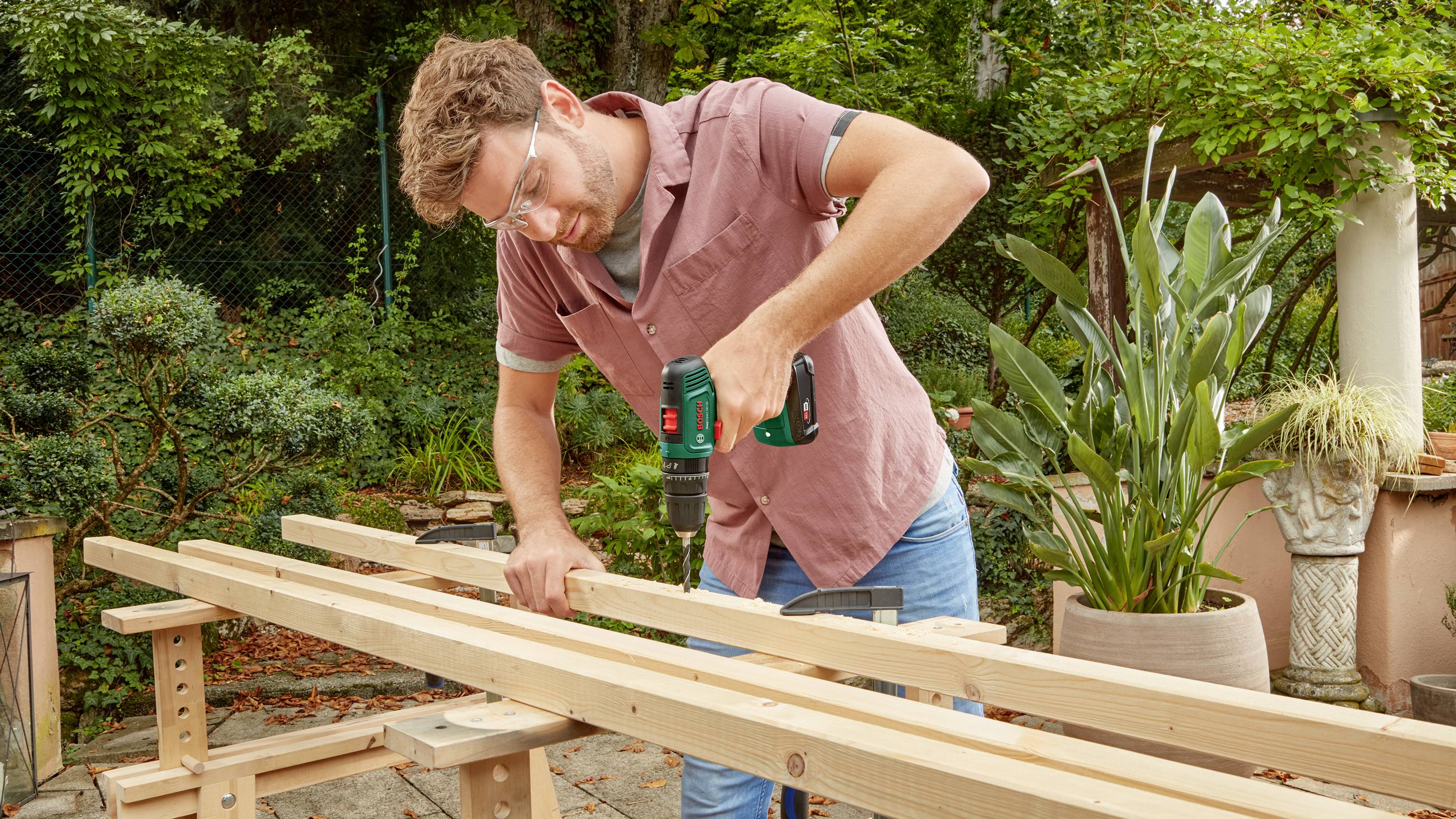 Ein Mann benutzt eine Bohrmaschine, um Holzlatten im Garten zusammenzuschrauben. Im Hintergrund sind Pflanzen und Terrakotta-Gefäße.