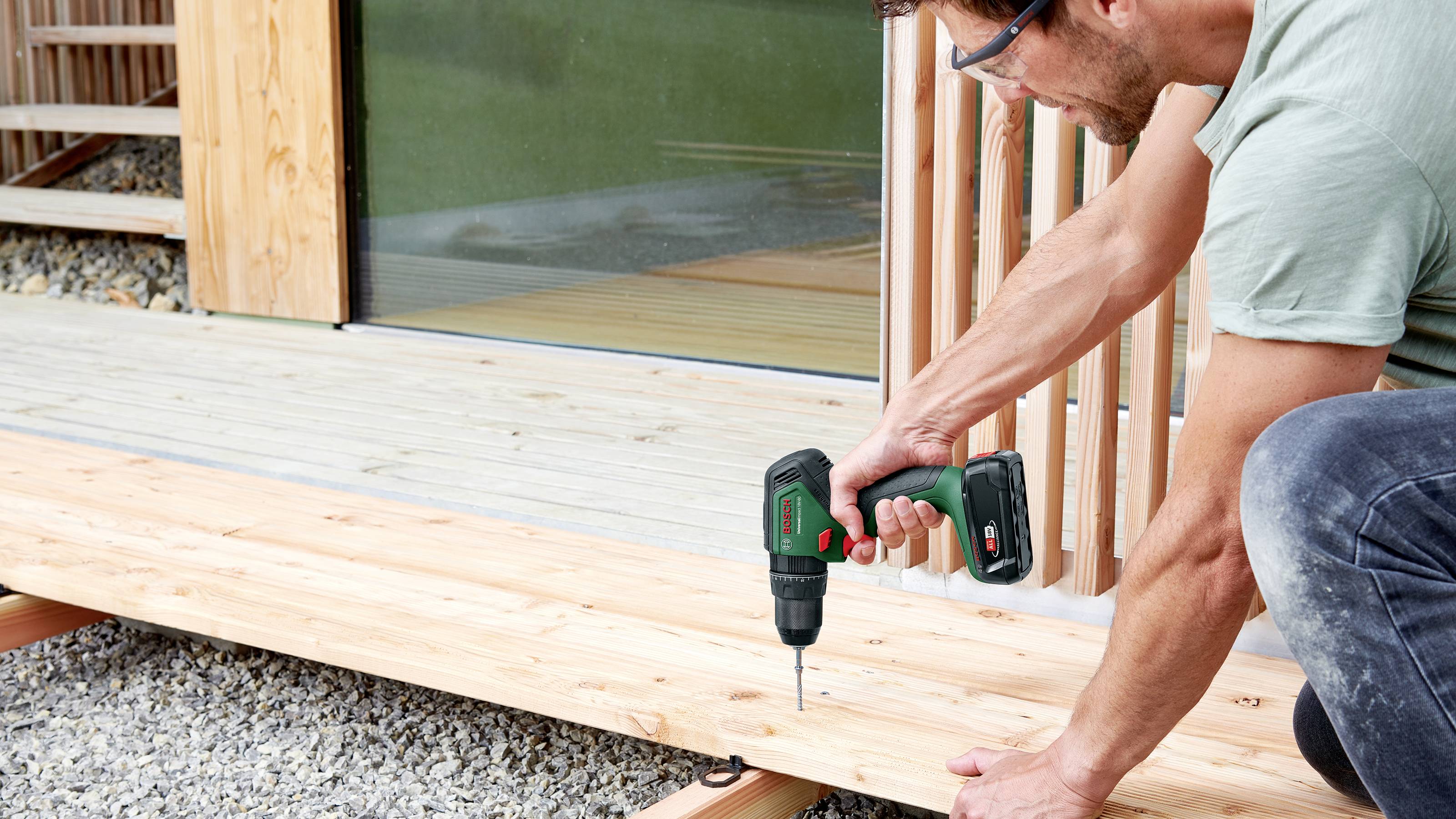 Ein Mann verwendet eine Bohrmaschine, um Holzplanken auf einer Terrasse zu befestigen. Er arbeitet konzentriert und trägt eine Schutzbrille.