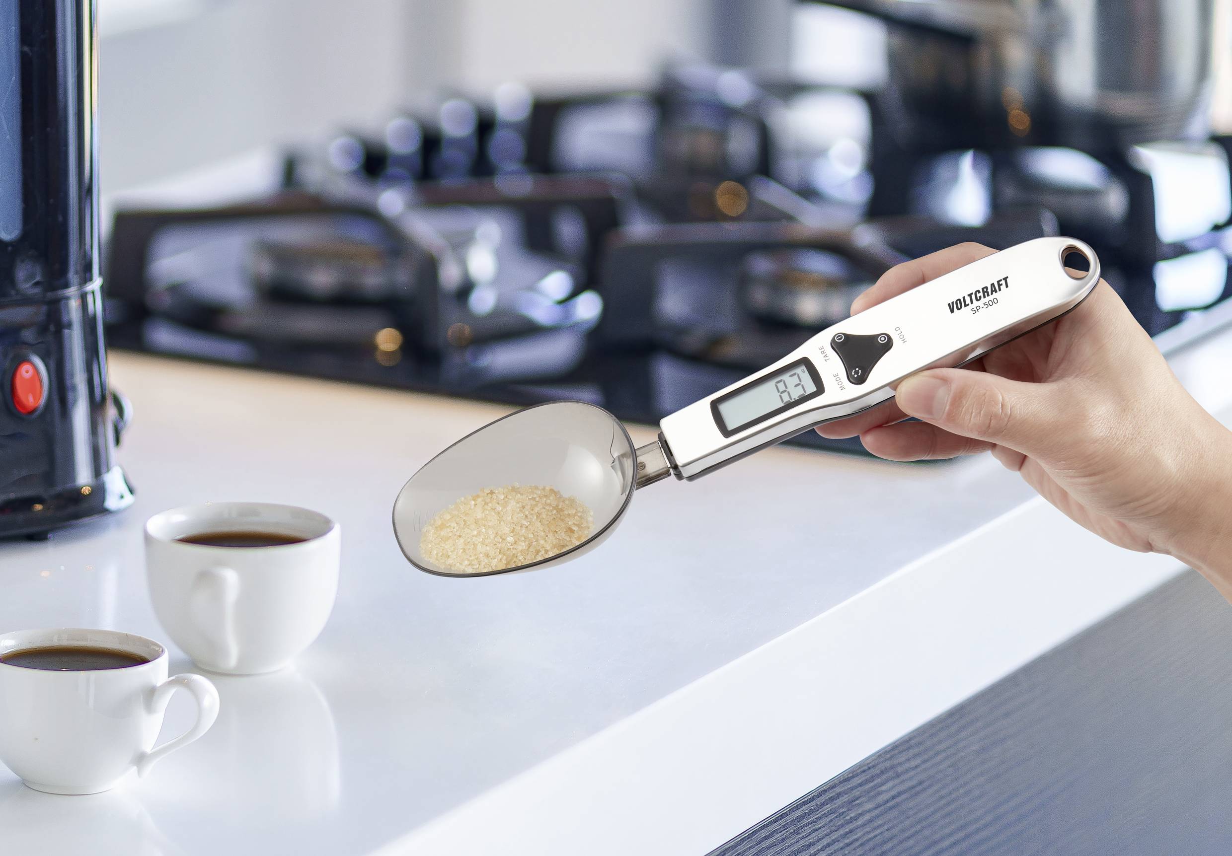 Person hält einen digitalen Löffel mit einem Display, das ein Gewicht anzeigt. Zwei Kaffeetassen stehen im Hintergrund auf einer Küchenarbeitsplatte.