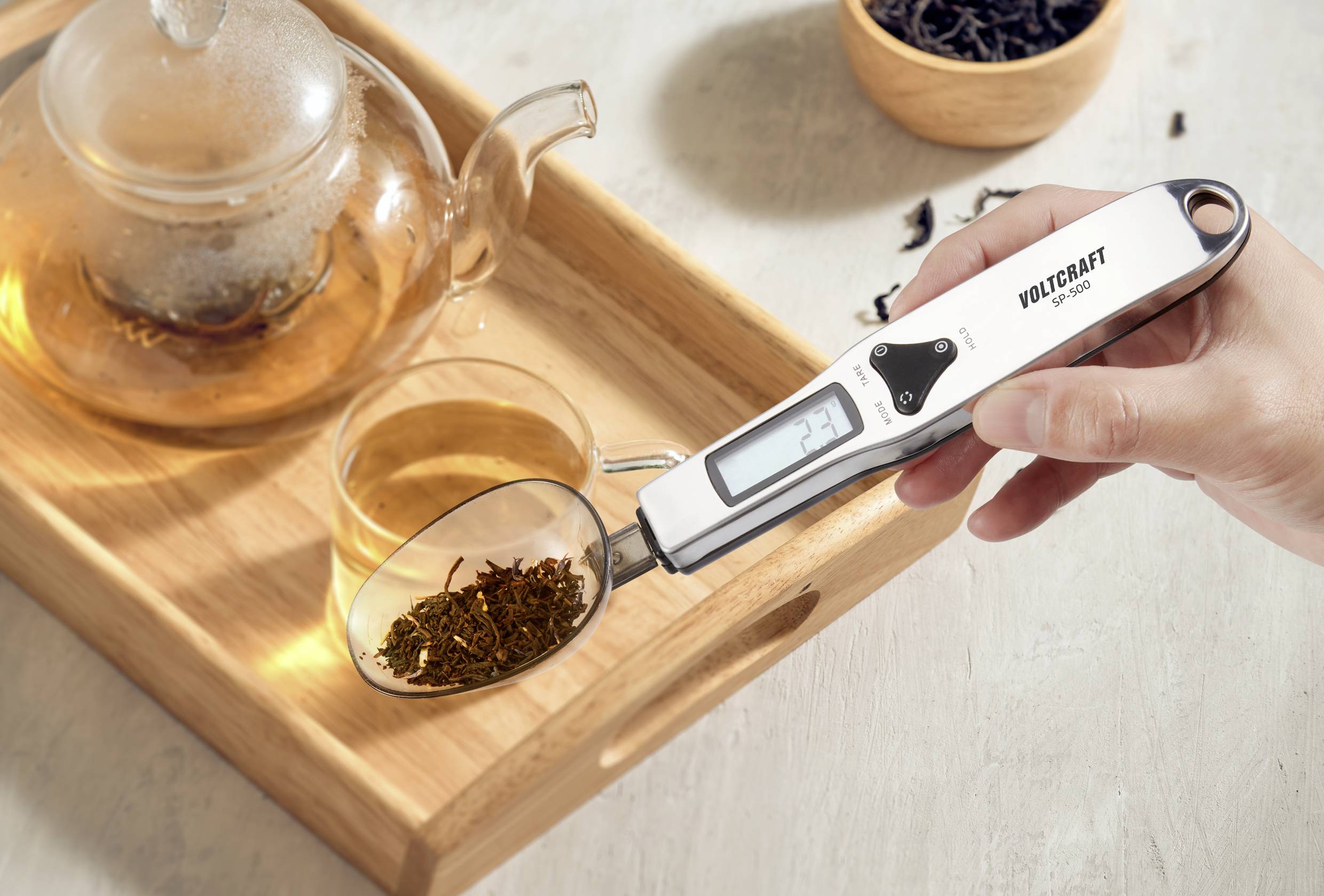 Eine Hand hält einen digitalen Löffelwaage mit Teeblättern, neben einer Glasteekanne und einer Tasse auf einem Tablett.