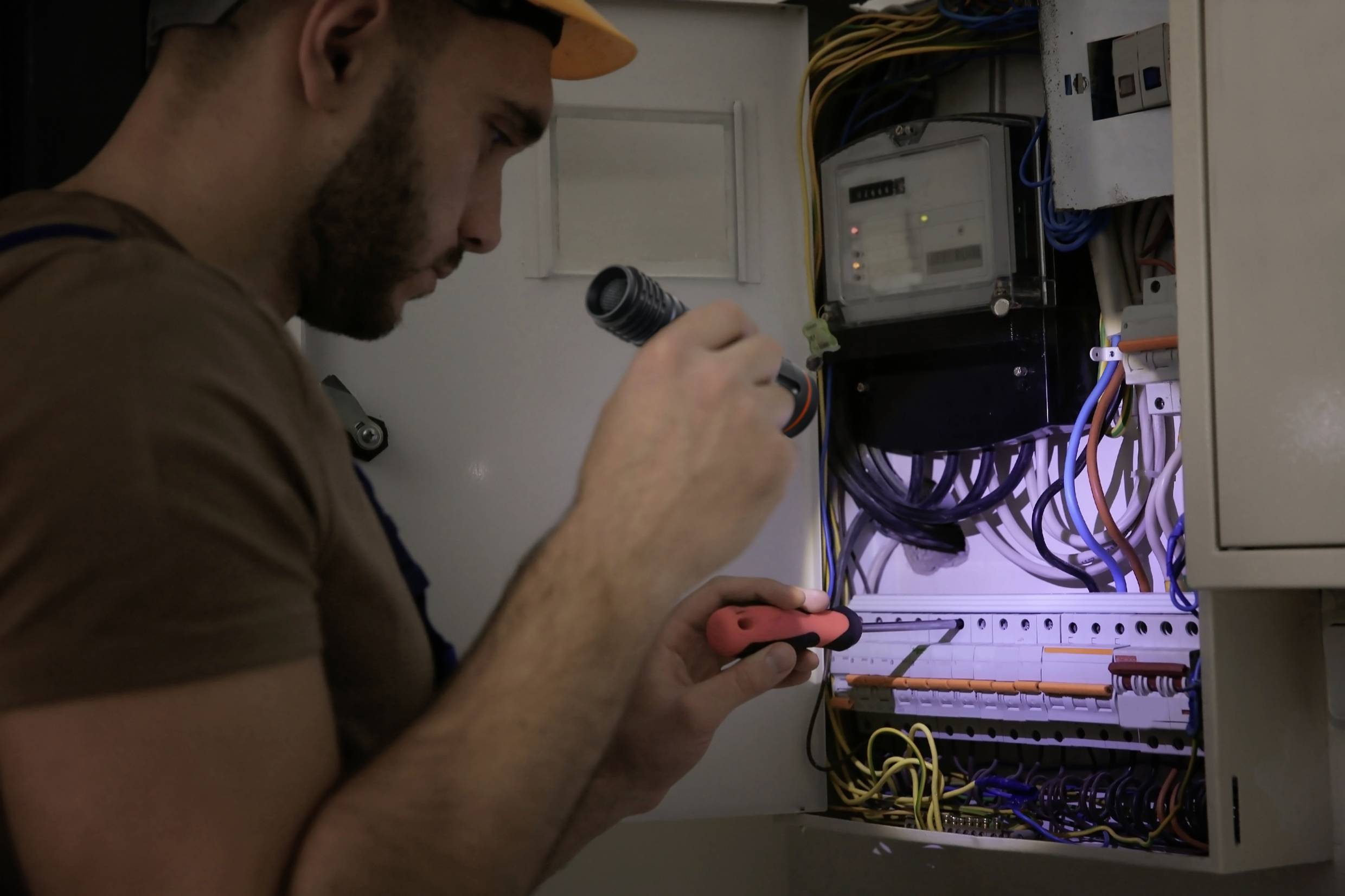Ein Elektriker arbeitet in einem Schaltschrank, hält eine Taschenlampe und benutzt einen Schraubendreher, um die Anschlüsse zu überprüfen.