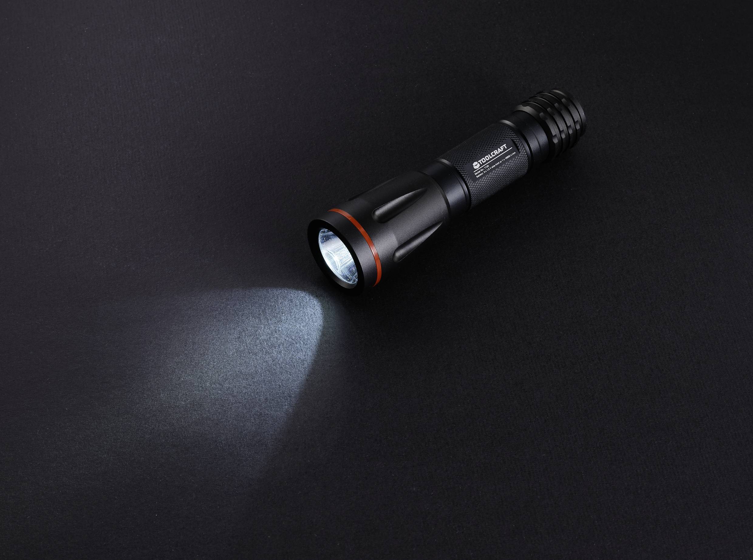 Eine schwarze Taschenlampe strahlt Licht auf eine dunkle Oberfläche. Sie liegt nach links gerichtet und ist das einzige Objekt im Bild.