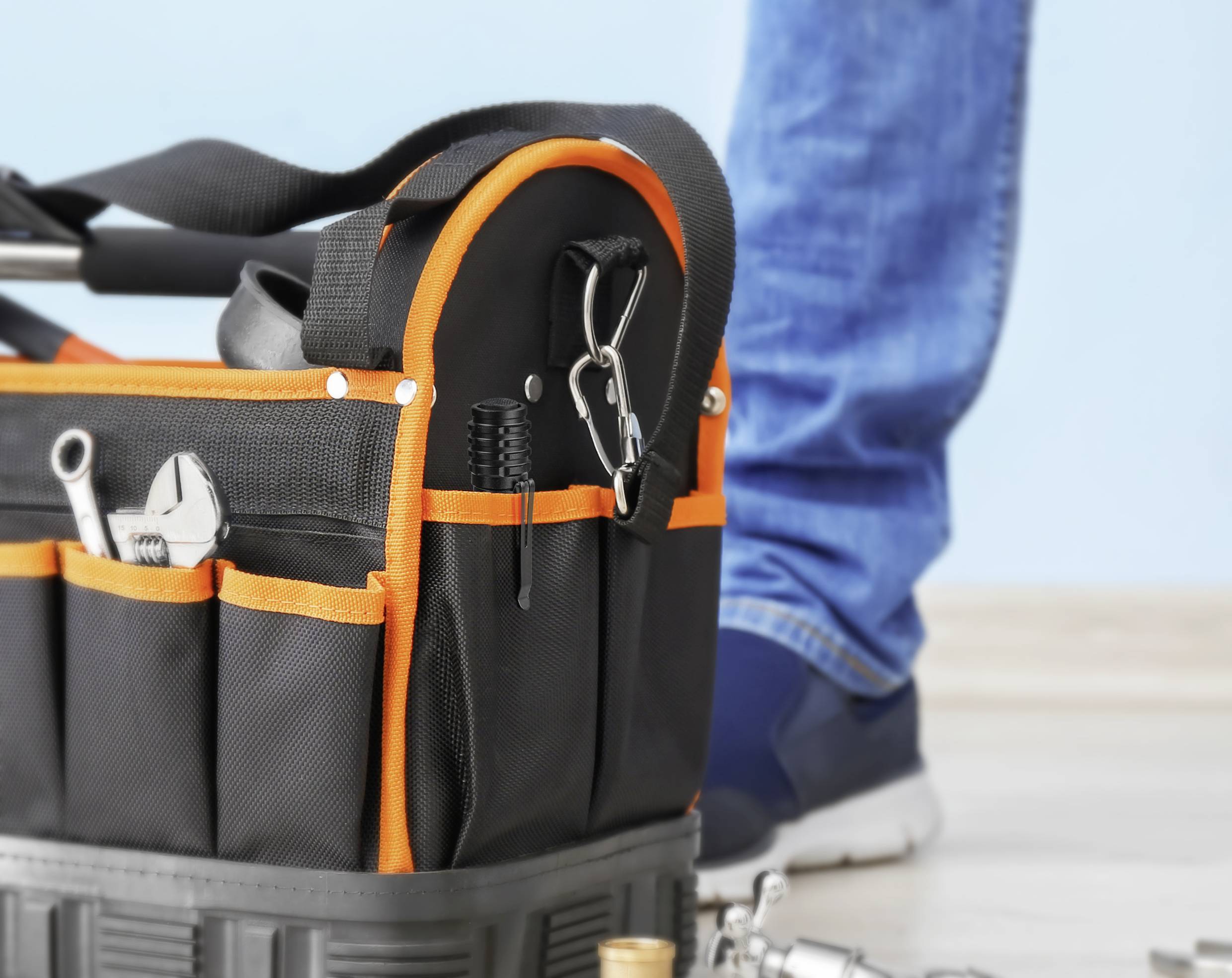 Eine schwarze Werkzeugtasche mit orangefarbenem Rand steht auf dem Boden. Daneben steht eine Person in blauer Jeans und Turnschuhen.