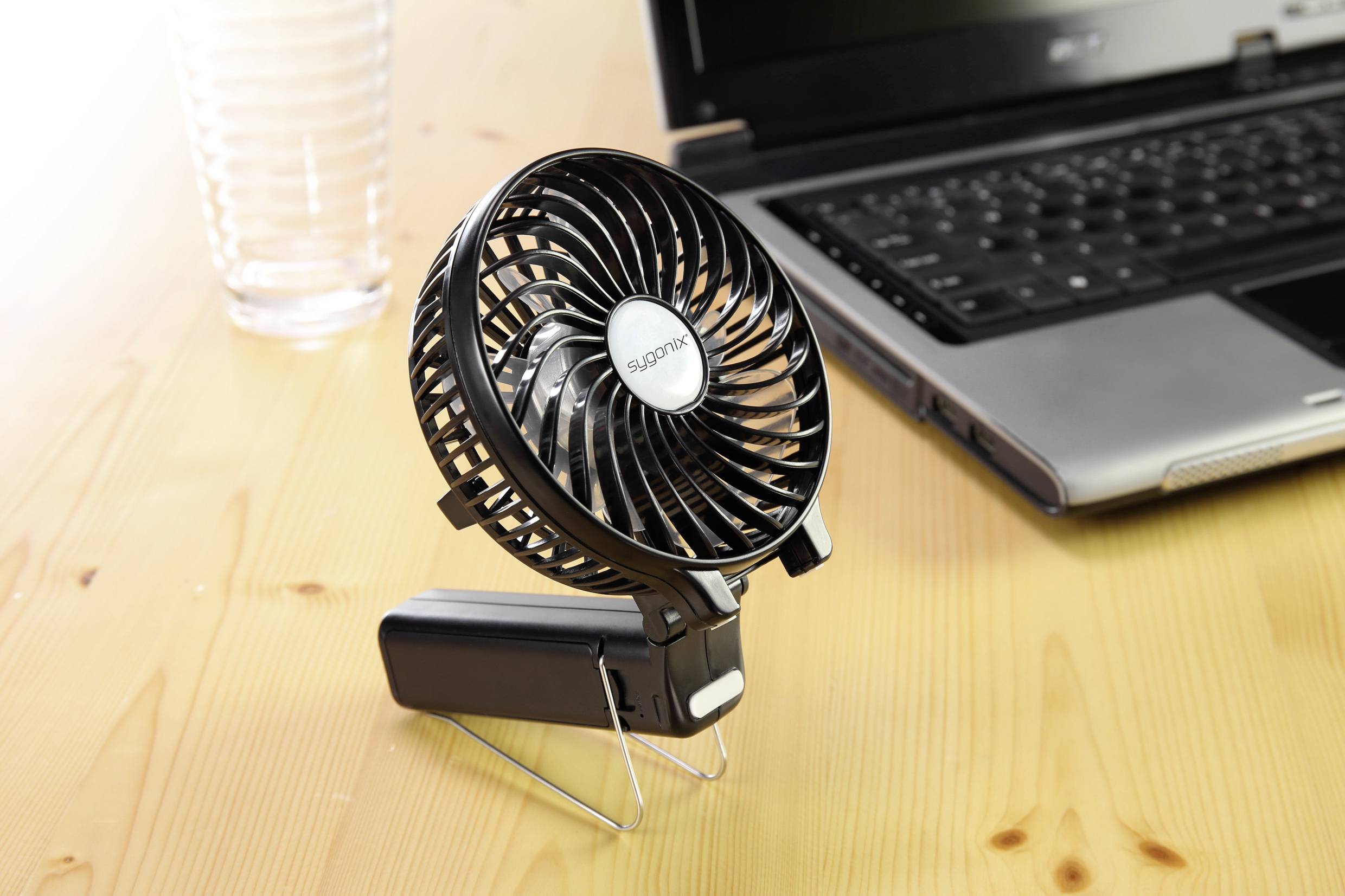 Ein kleiner schwarzer Tischventilator steht auf einem Holztisch neben einem Laptop und einem leeren Glas.