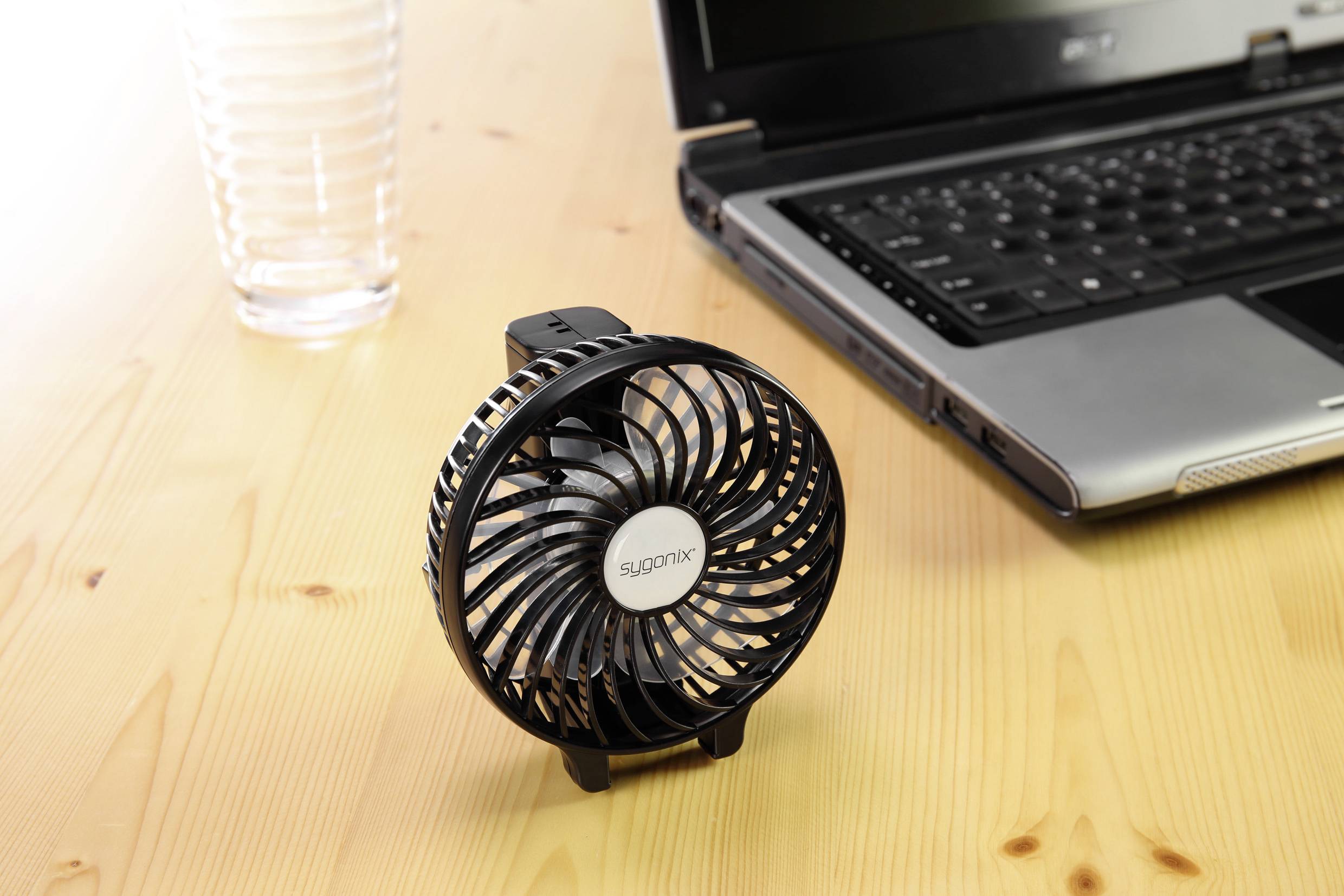 Kleiner schwarzer Tischventilator neben einem Laptop und einem Glas auf einem Holztisch.