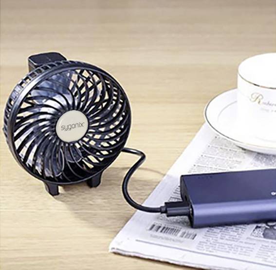 Ein kleiner Ventilator, angeschlossen an eine Powerbank, steht auf einem Tisch neben einer Kaffeetasse und einer Zeitung.