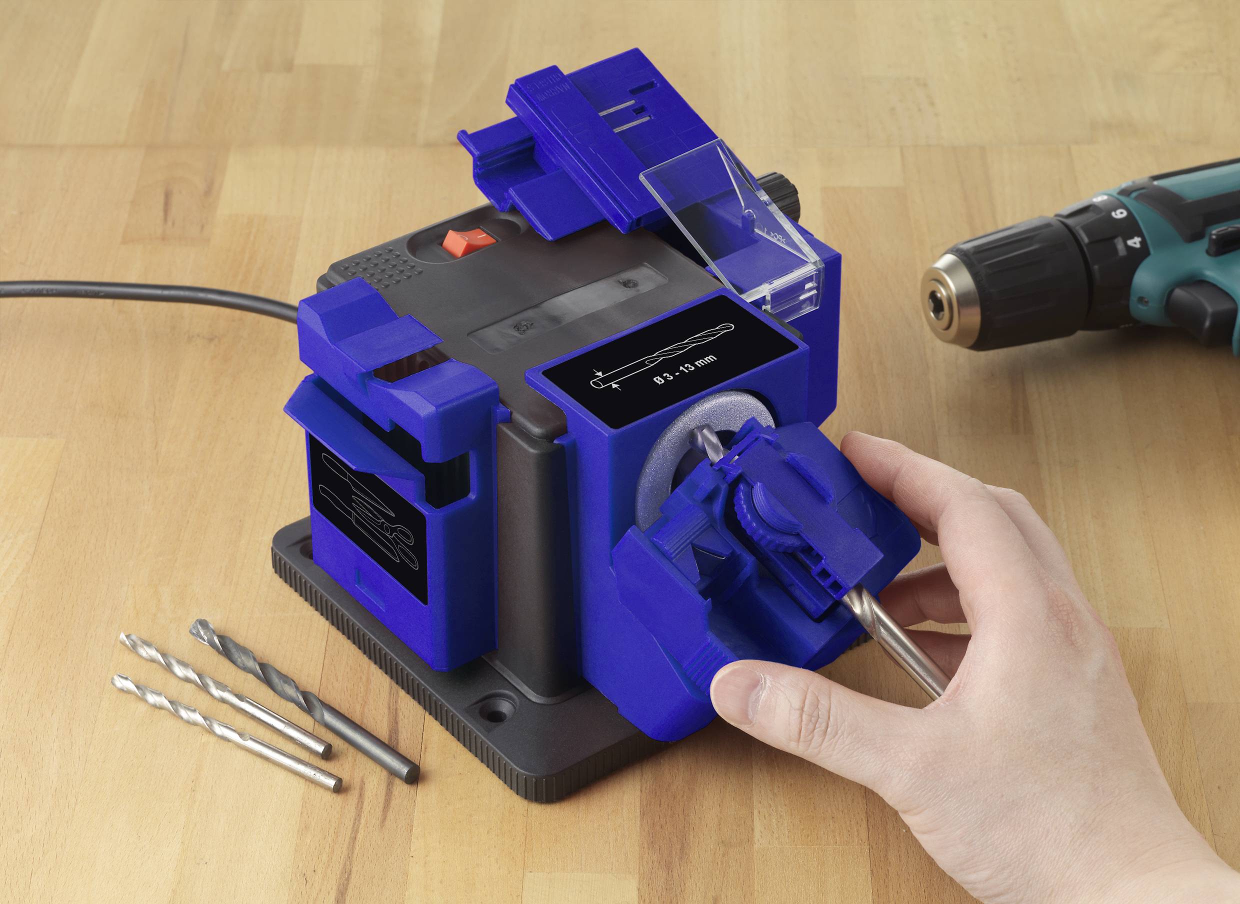 Eine Person verwendet ein blaues Schleifgerät auf einem Holztisch, um einen Bohrer zu schärfen. Daneben liegen drei weitere Bohrer und eine Bohrmaschine.