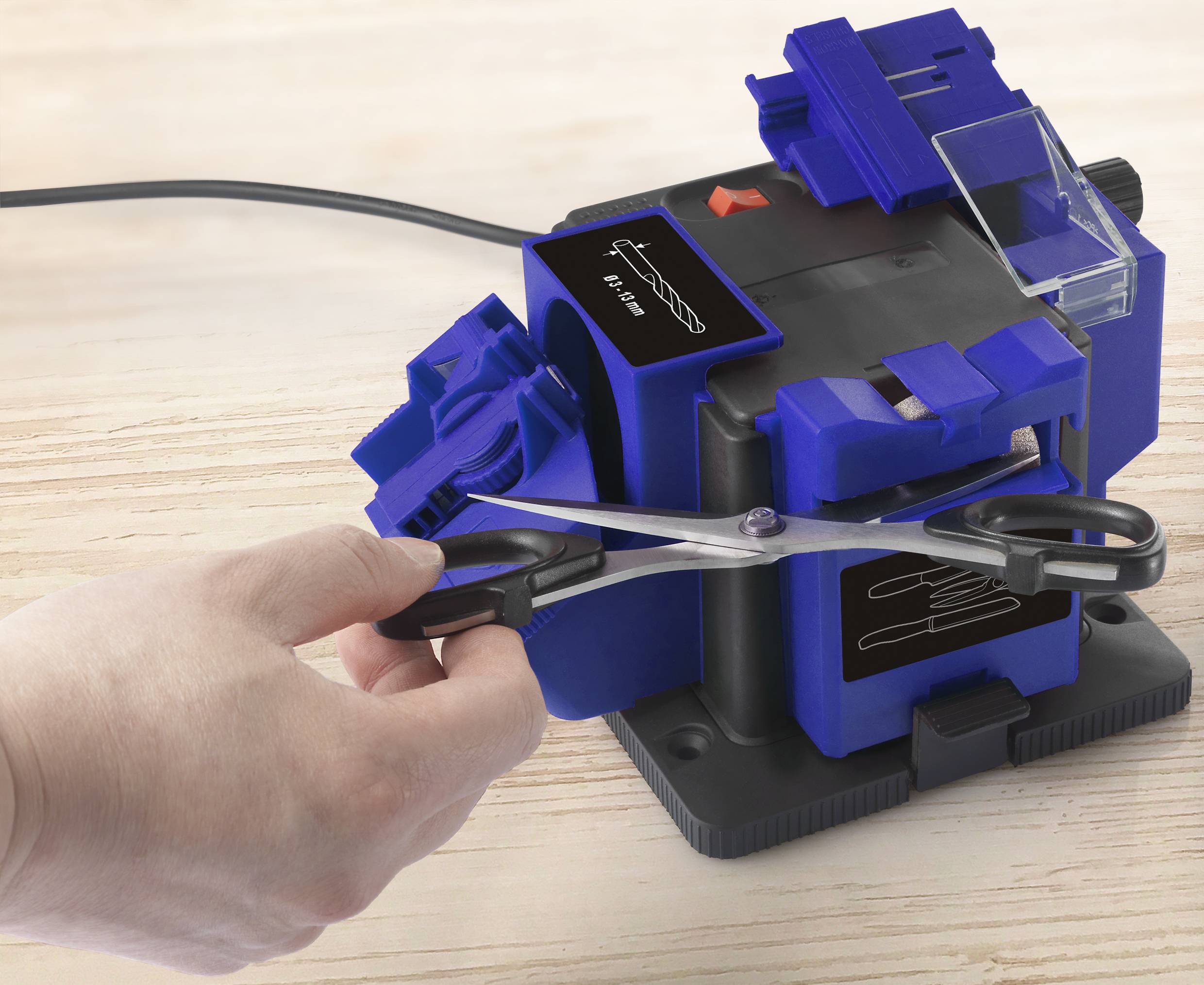 Eine Hand schärft eine Schere mit einem blau-schwarzen Schärfgerät. Das Gerät steht auf einer Holzoberfläche.