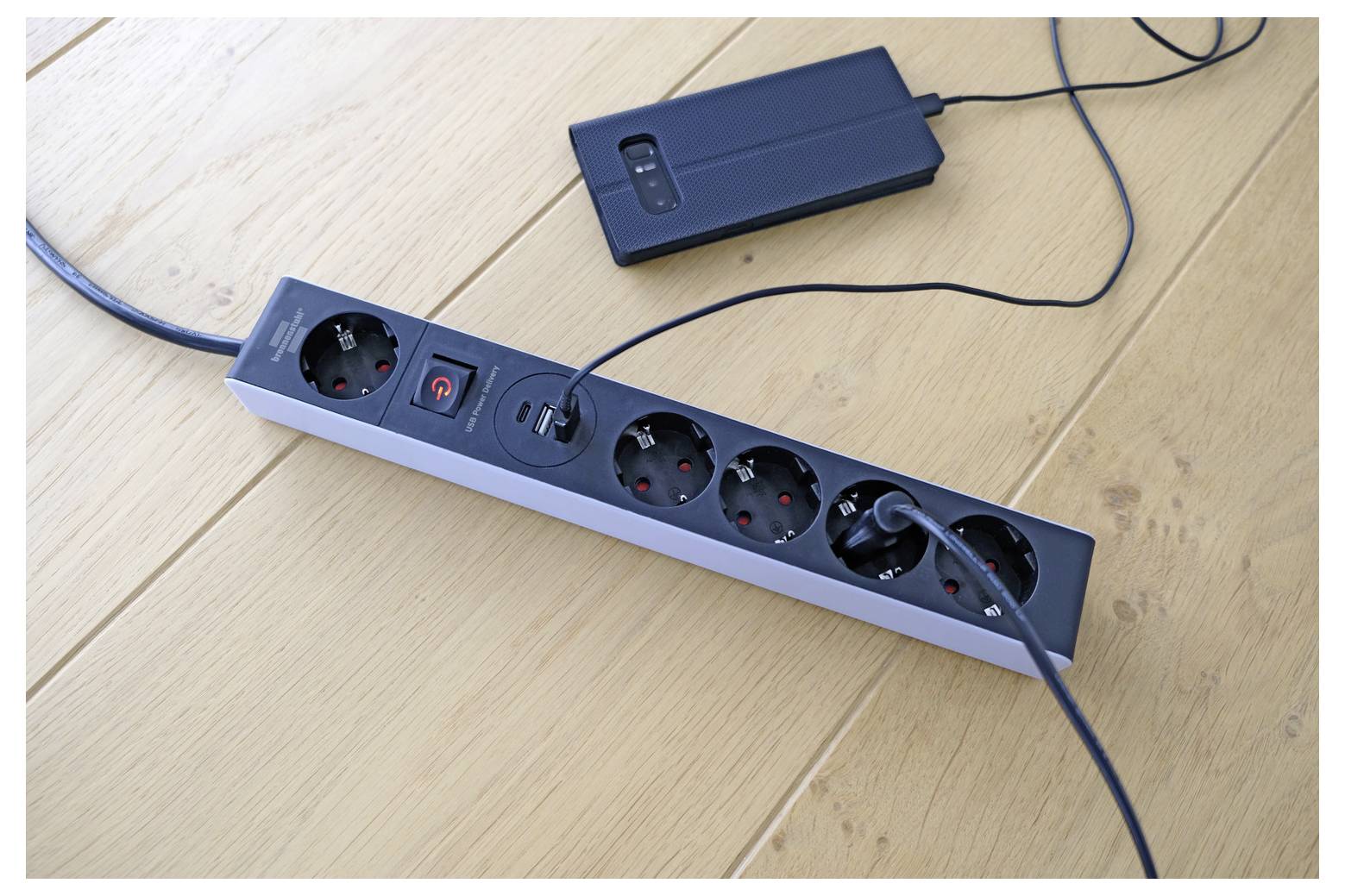 Ein schwarze Steckerleiste mit mehreren Steckdosen und eingesteckten Kabeln liegt auf einem Holzboden neben einer tragbaren Powerbank oder einem Ladegerät.