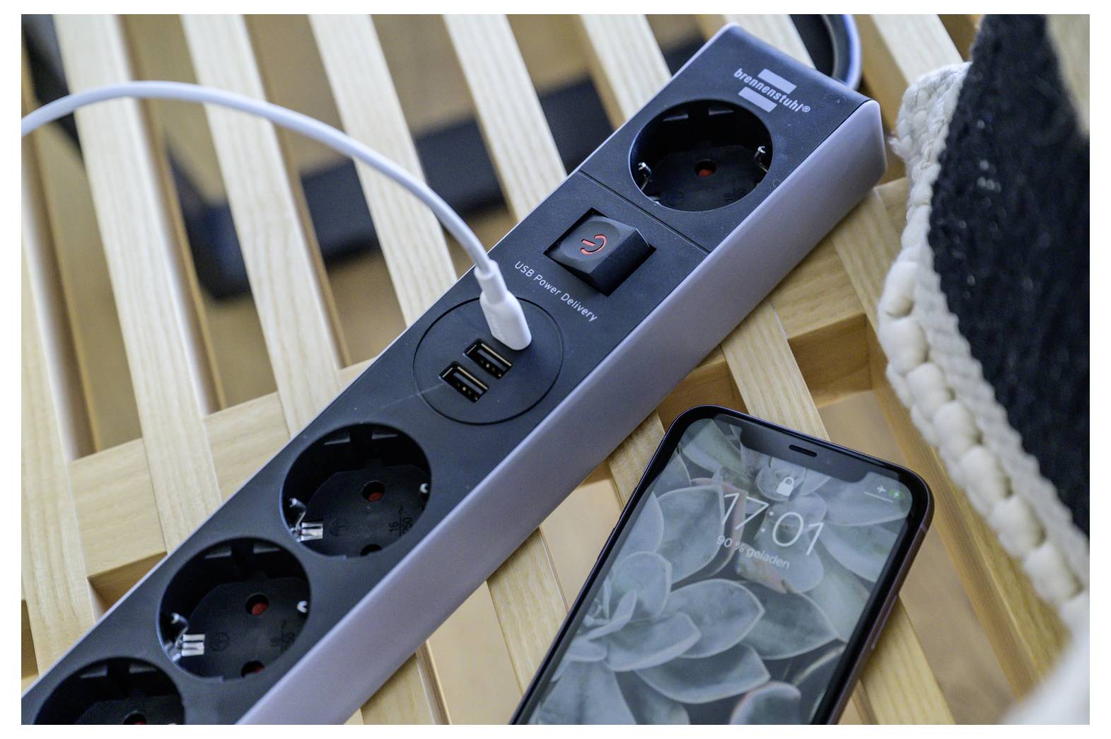 Steckerleiste mit einem USB-Ladekabel daneben angeschlossen neben einem Smartphone auf einer Holzlattenfläche.