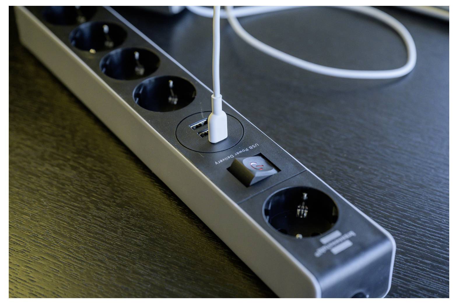 Nahaufnahme einer schwarzen Steckerleiste auf einem Schreibtisch, mit einem USB-Kabel, das in den integrierten Ladeanschluss eingesteckt ist, und einem sichtbaren Ein-/Ausschalter.