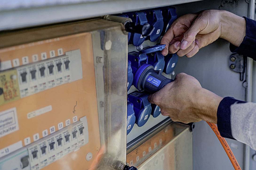 Eine Person steckt einen blauen Stecker in eine Reihe von Steckdosen in einem elektrischen Schaltschrank.