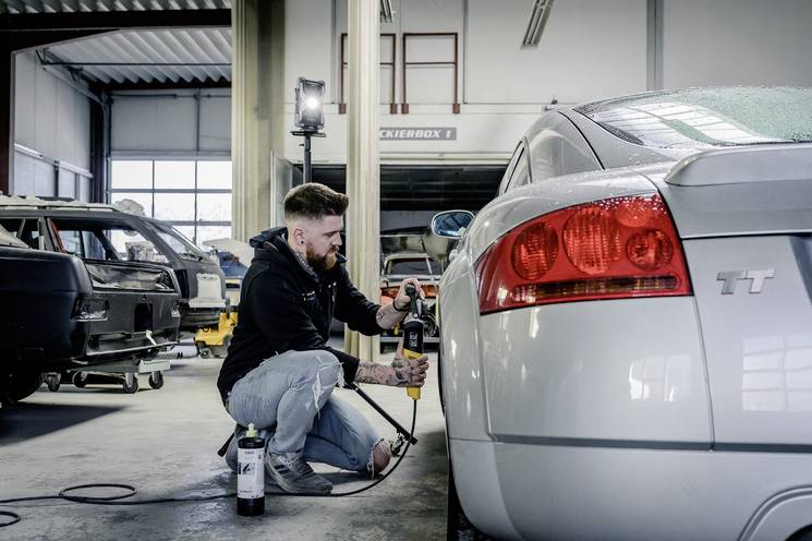 Ein Mann arbeitet in einer Autowerkstatt an einem silbernen Sportwagen. Er benutzt ein Werkzeug, um am Fahrzeug zu arbeiten. Im Hintergrund stehen andere Autos.