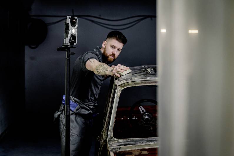 Ein Mann poliert mit einem Tuch ein altes Auto in einer Werkstatt. Er trägt ein Tattoo auf dem Arm. Im Hintergrund steht ein Arbeitslicht.