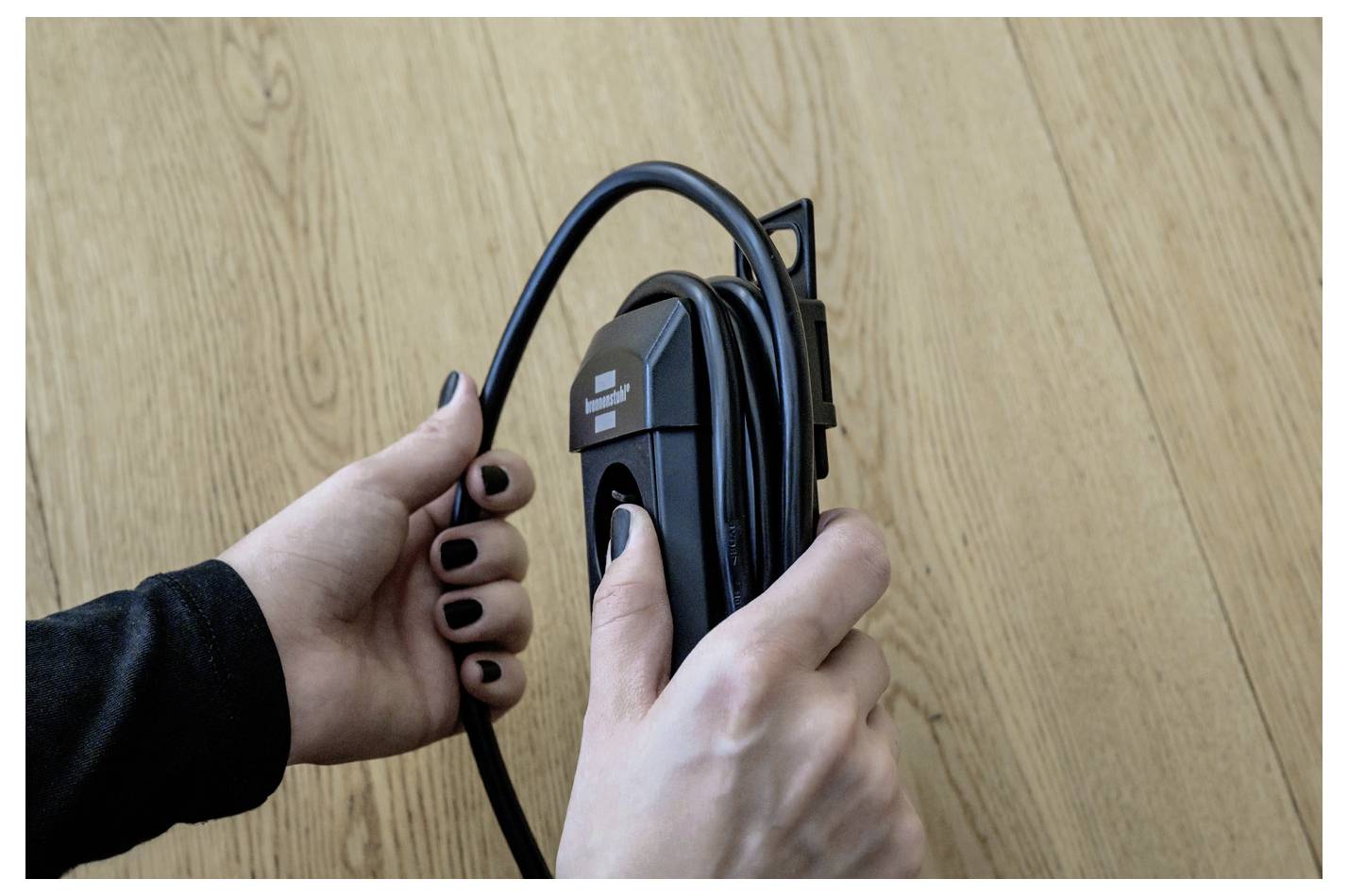 Hände halten und wickeln ein schwarzes Ladekabel um eine wandmontierte Halterung auf einer Holzoberfläche.