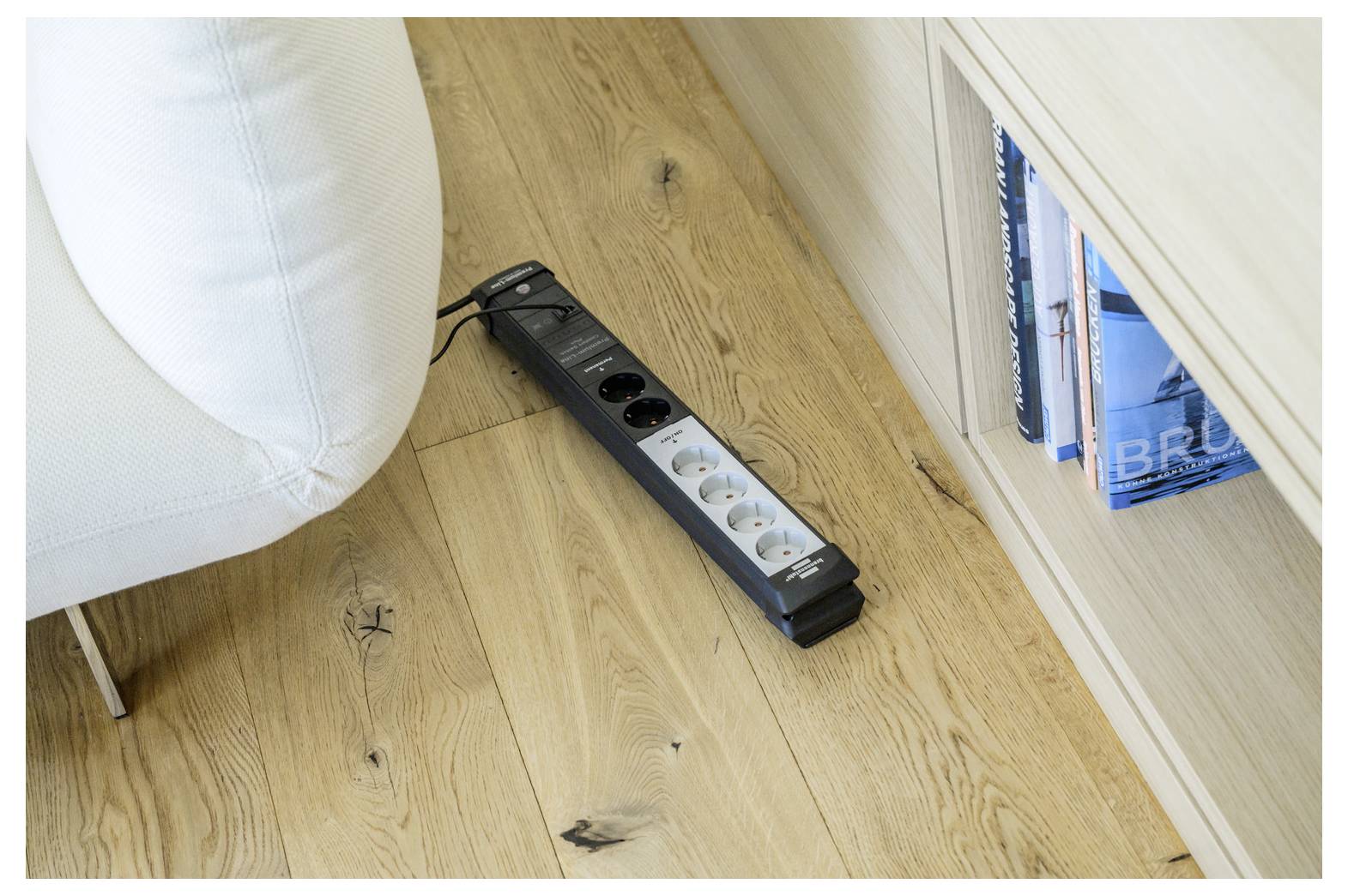 Schwarze Steckerleiste mit mehreren Steckdosen liegt auf einem Holzboden neben einem weißen Sofa und einem Bücherregal.