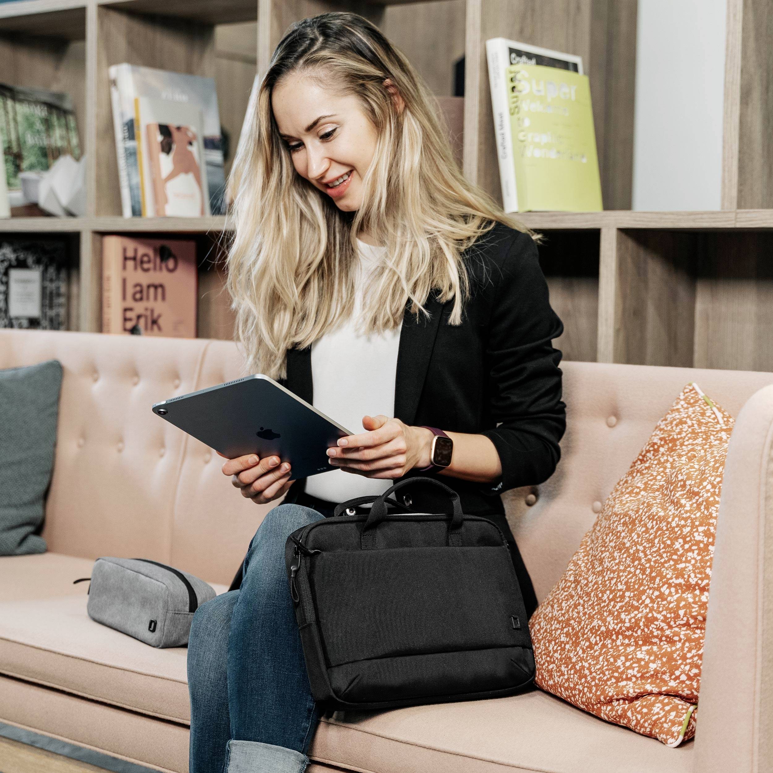 Eine lächelnde Frau sitzt auf einem Sofa und hält ein Tablet. Im Hintergrund sind Bücherregale sichtbar. Eine Tasche liegt neben ihr.