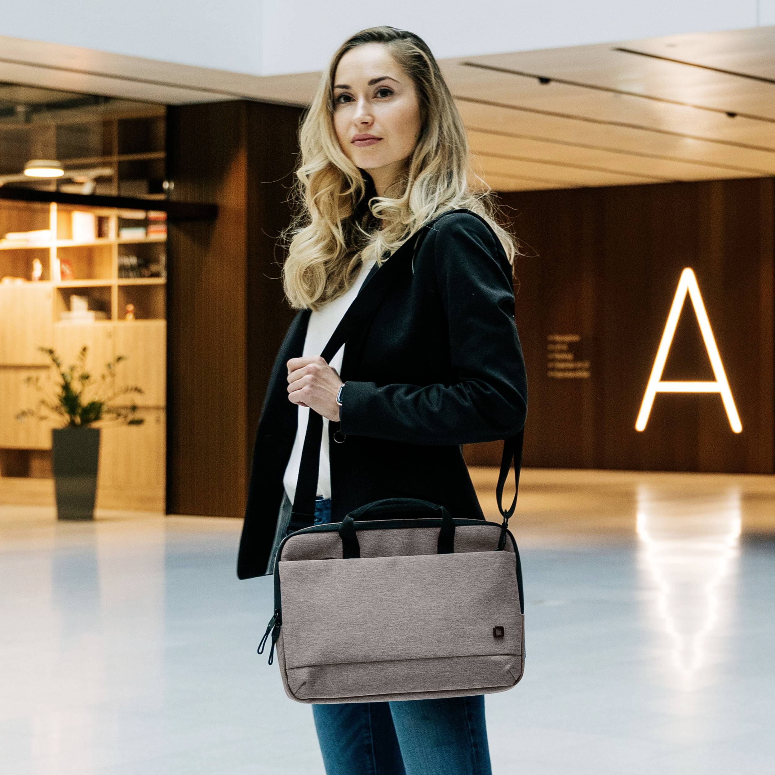 Person in einem modernen Gebäude, trägt formelle Kleidung und hält eine Laptoptasche. Hintergrund hat stilvolle Holzwände und Beleuchtung.
