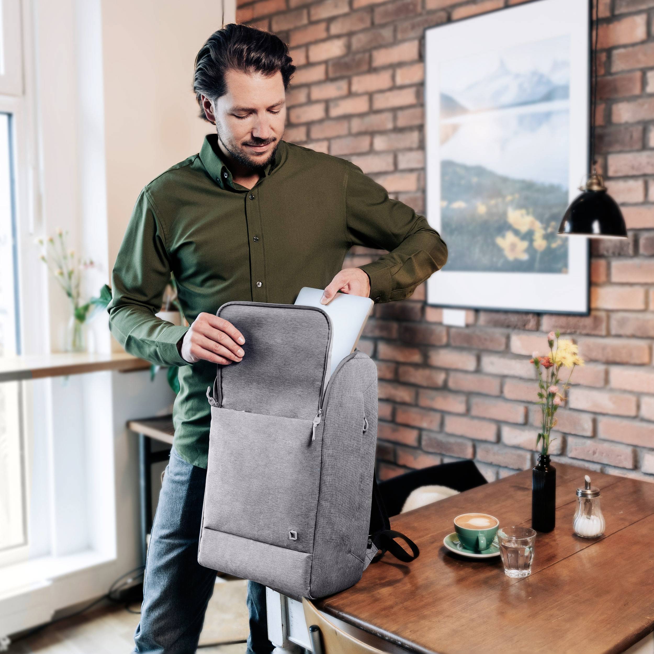 Ein Mann packt einen Laptop aus einem Rucksack in einem Café aus. Auf dem Tisch stehen eine Tasse Kaffee und ein Teller.