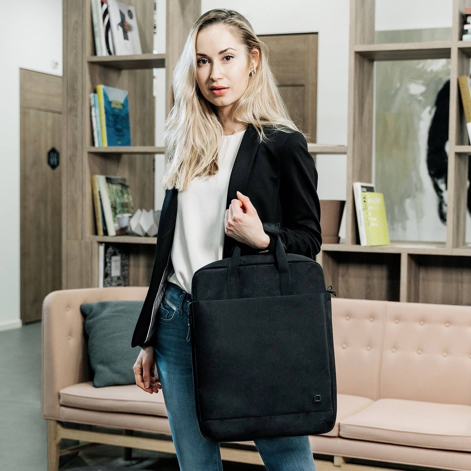 Eine Frau steht in einem modernen Büro, hält eine schwarze Tasche, trägt einen Blazer und Jeans. Im Hintergrund sind Regale mit Büchern.