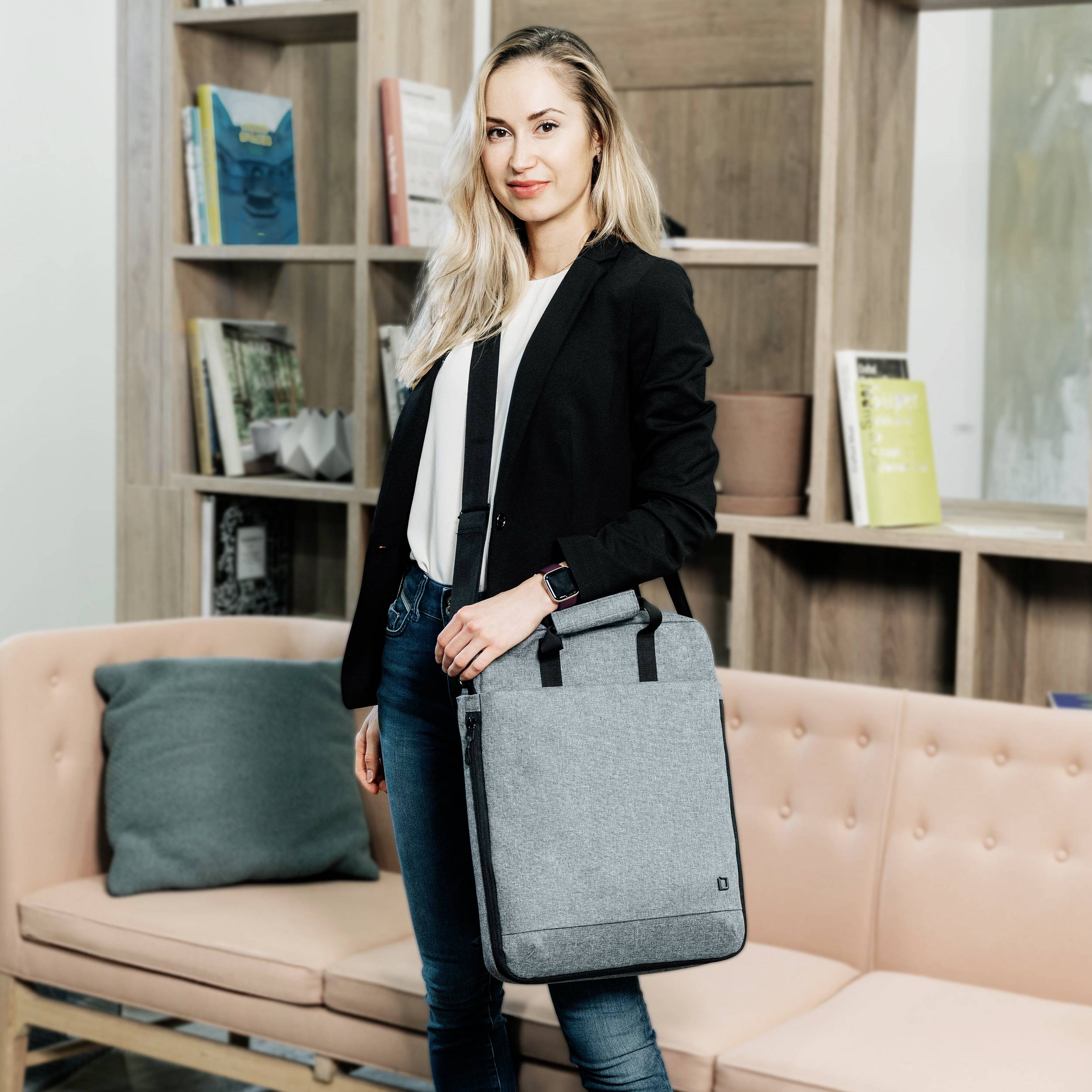 Eine Person steht in einem modern eingerichteten Raum vor einem Bücherregal. Sie trägt ein schwarzes Sakko und hält eine graue Tasche in der Hand.