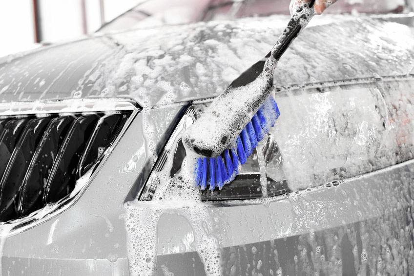 Eine Hand wäscht mit einer blauen Bürste den Vorderteil eines silbernen Autos, das mit Seifenwasser bedeckt ist.