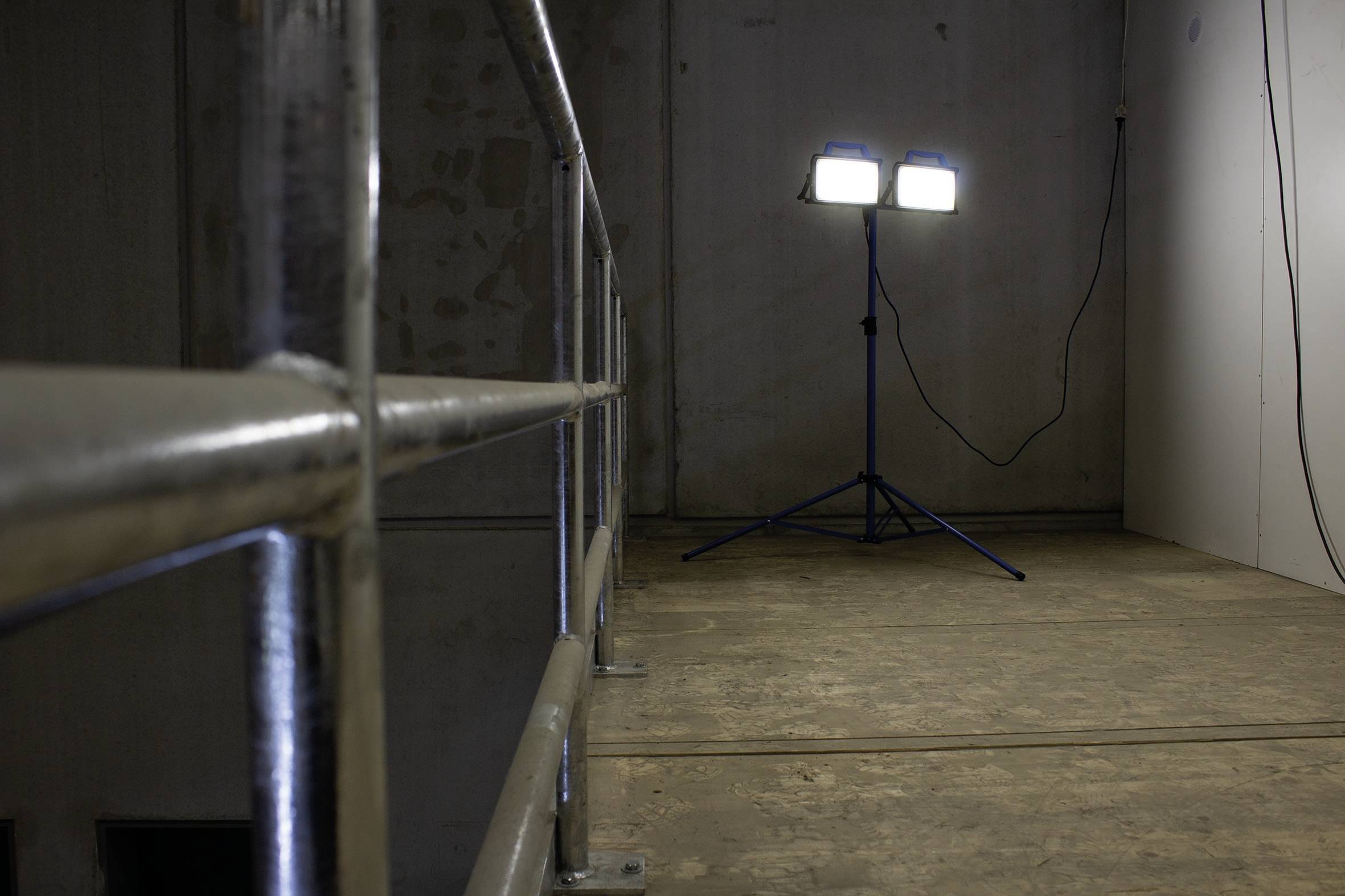 Baugerüst mit Sicherheitsgeländer in einem schwach beleuchteten Raum. Zwei helle Baustrahler stehen in der Ecke.