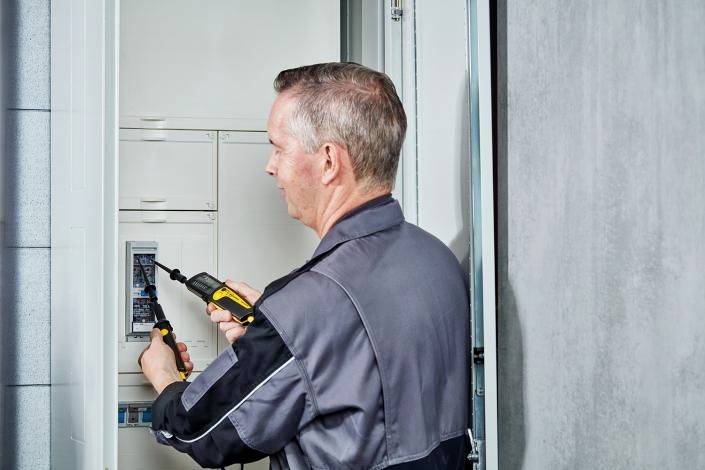 Ein Elektriker in Arbeitsuniform arbeitet mit einem Schraubendreher an einem geöffneten Stromverteilerkasten in einem Innenraum.