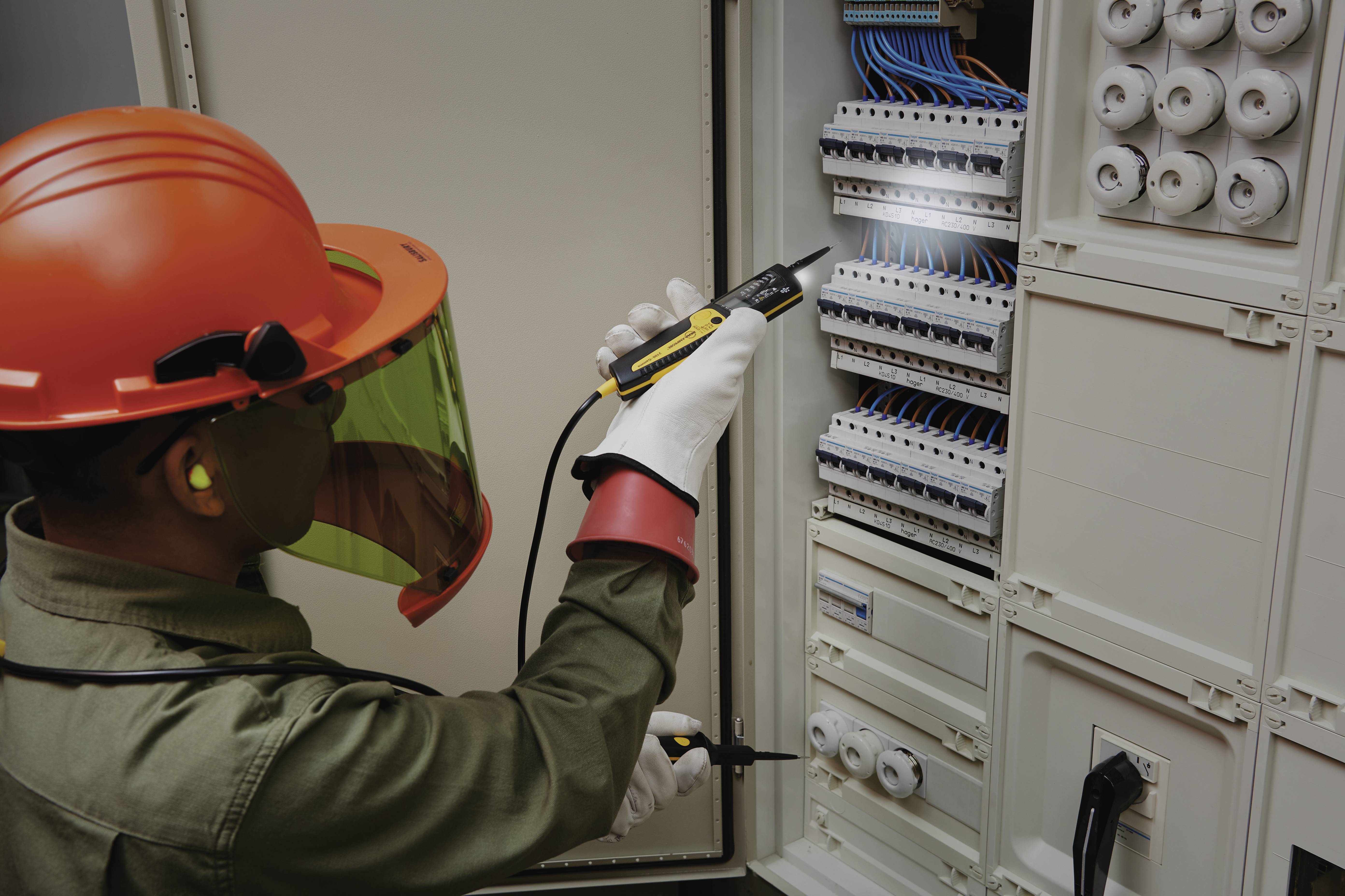 Ein Elektriker mit Schutzhelm und -brille arbeitet mit einem Testgerät an einem offenen Schaltschrank voller Kabel und Sicherungen.