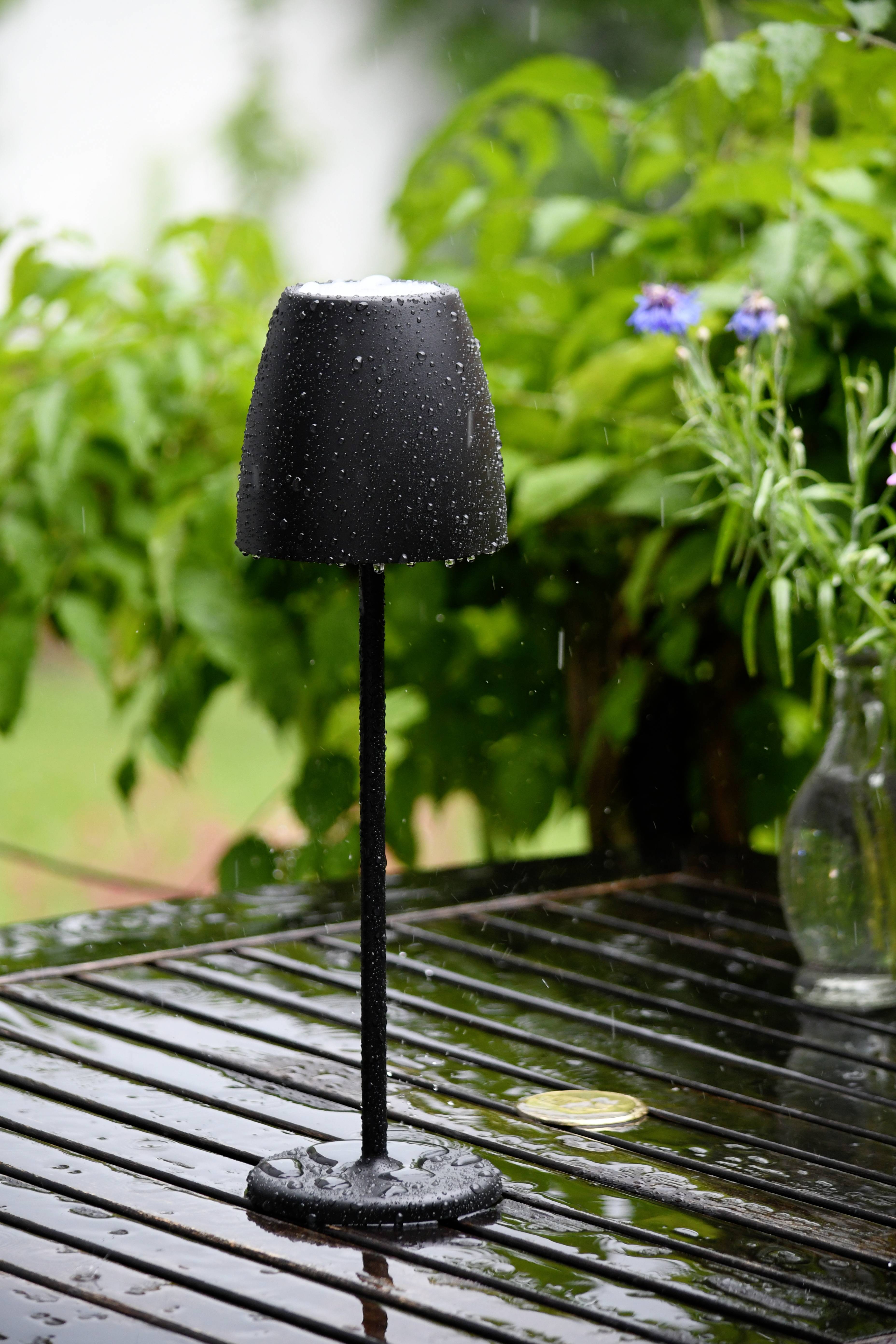 Eine schwarze Lampe steht auf einem nassen Holztisch im Freien. Im Hintergrund sind grüne Pflanzen und blühende Blumen zu sehen.