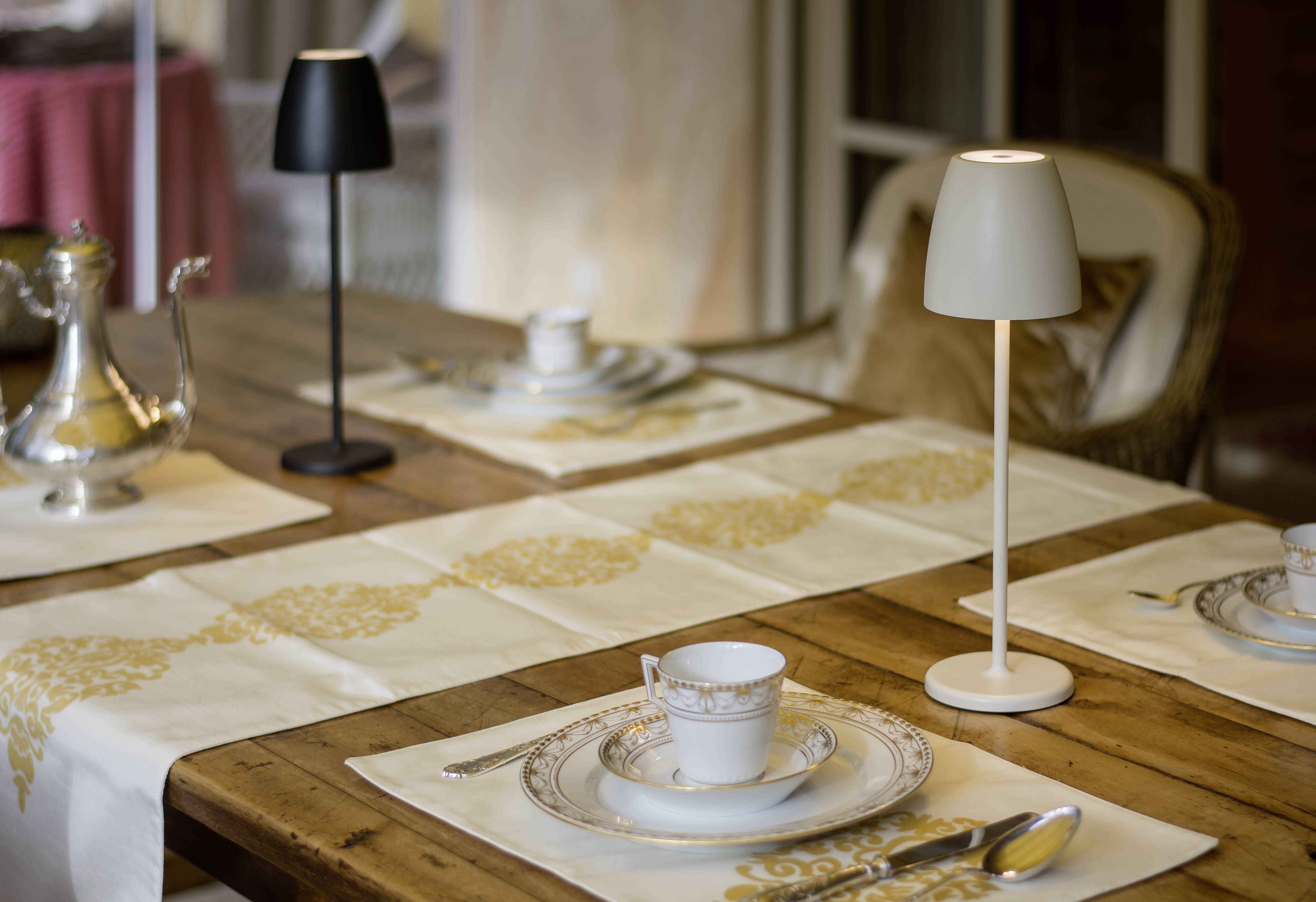 Eleganter Esstisch mit Tischdecken, gedeckt mit Geschirr, Tassen und Teekanne; zwei Tischlampen als Beleuchtung.