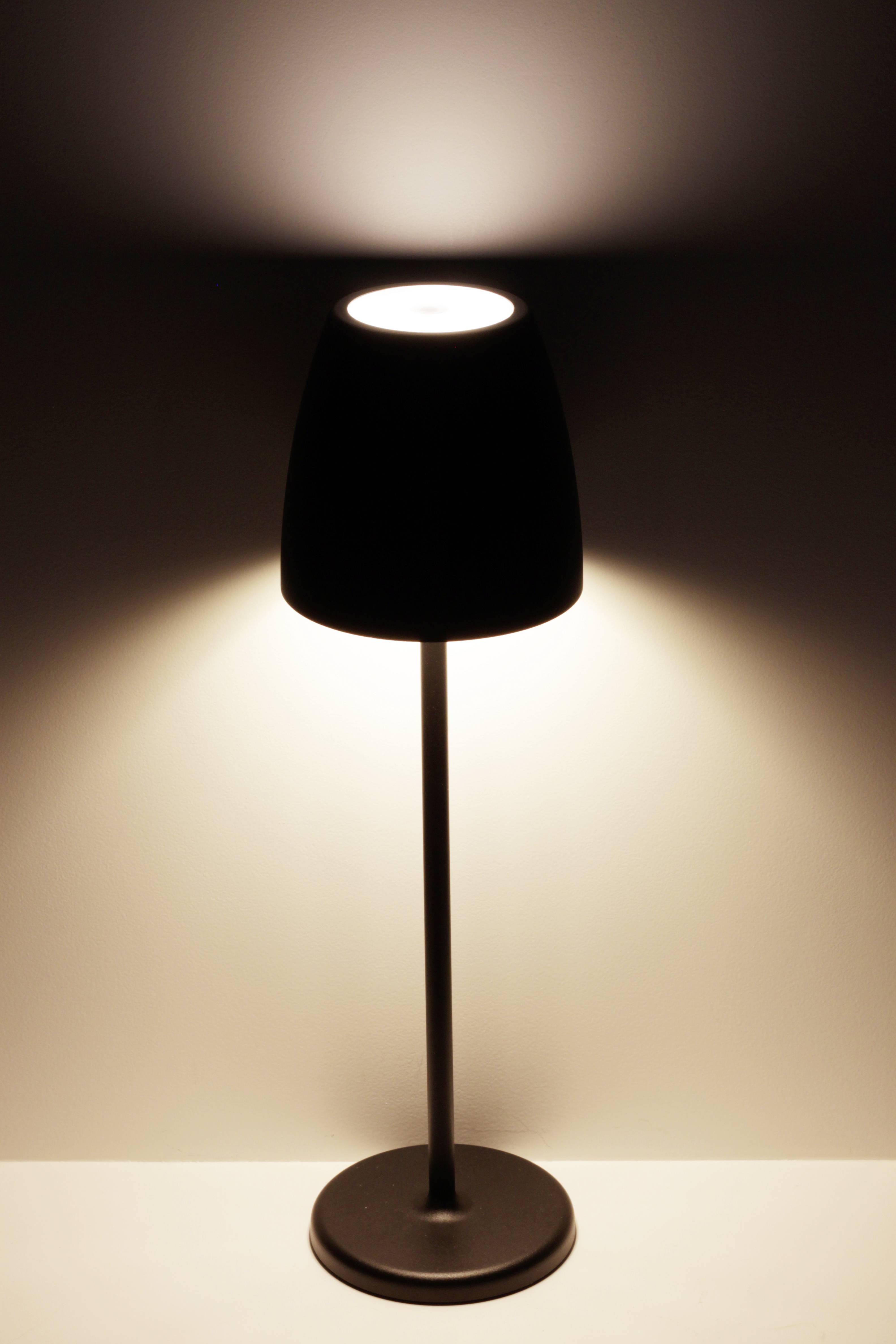 Eine minimalistische, schwarze Tischlampe auf einem weißen Tisch vor einer weißen Wand leuchtet nach oben und unten.