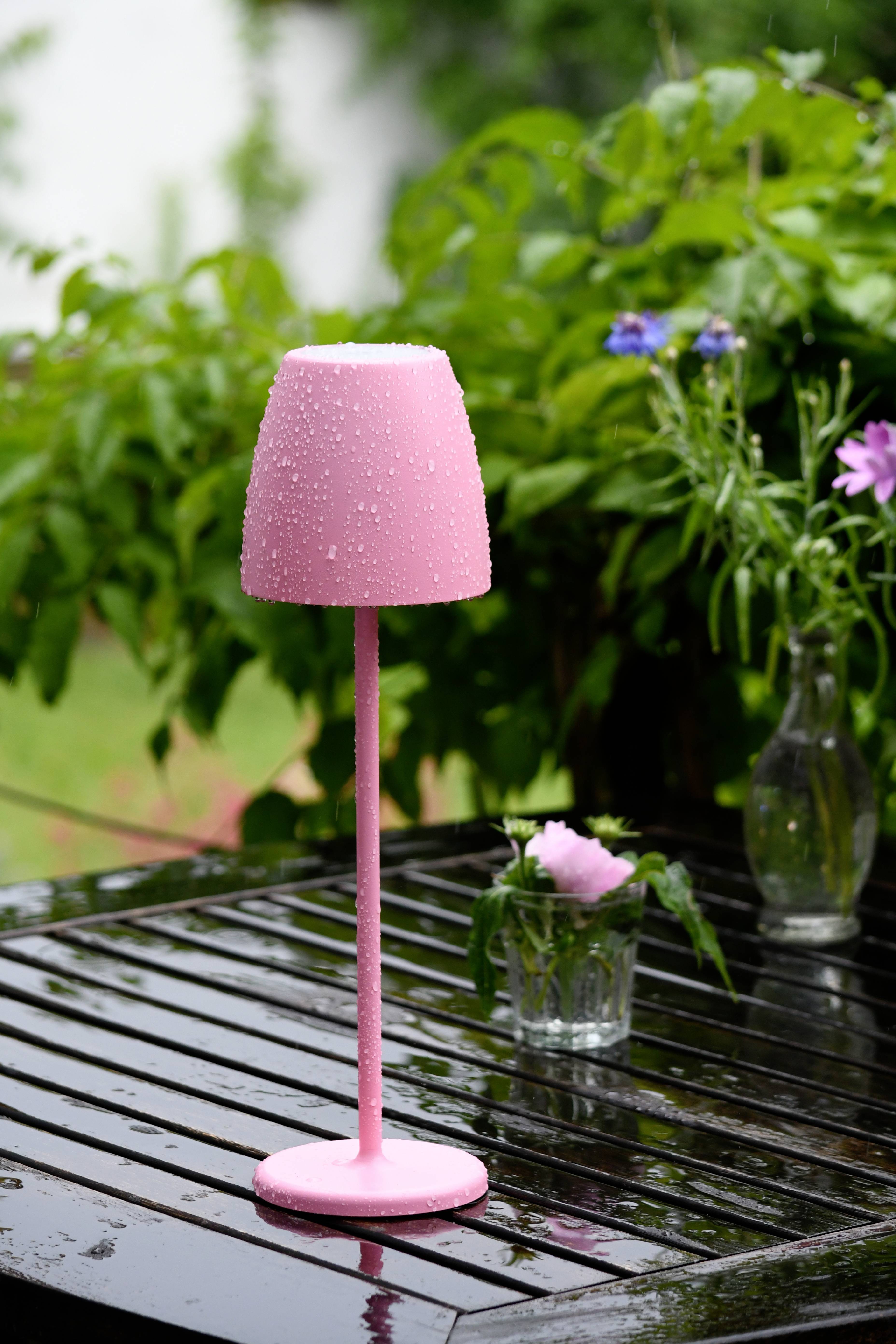 Eine rosa Lampe mit Wassertropfen steht auf einem nassen Holztisch im Freien. Im Hintergrund sind grüne Pflanzen und Blumen.
