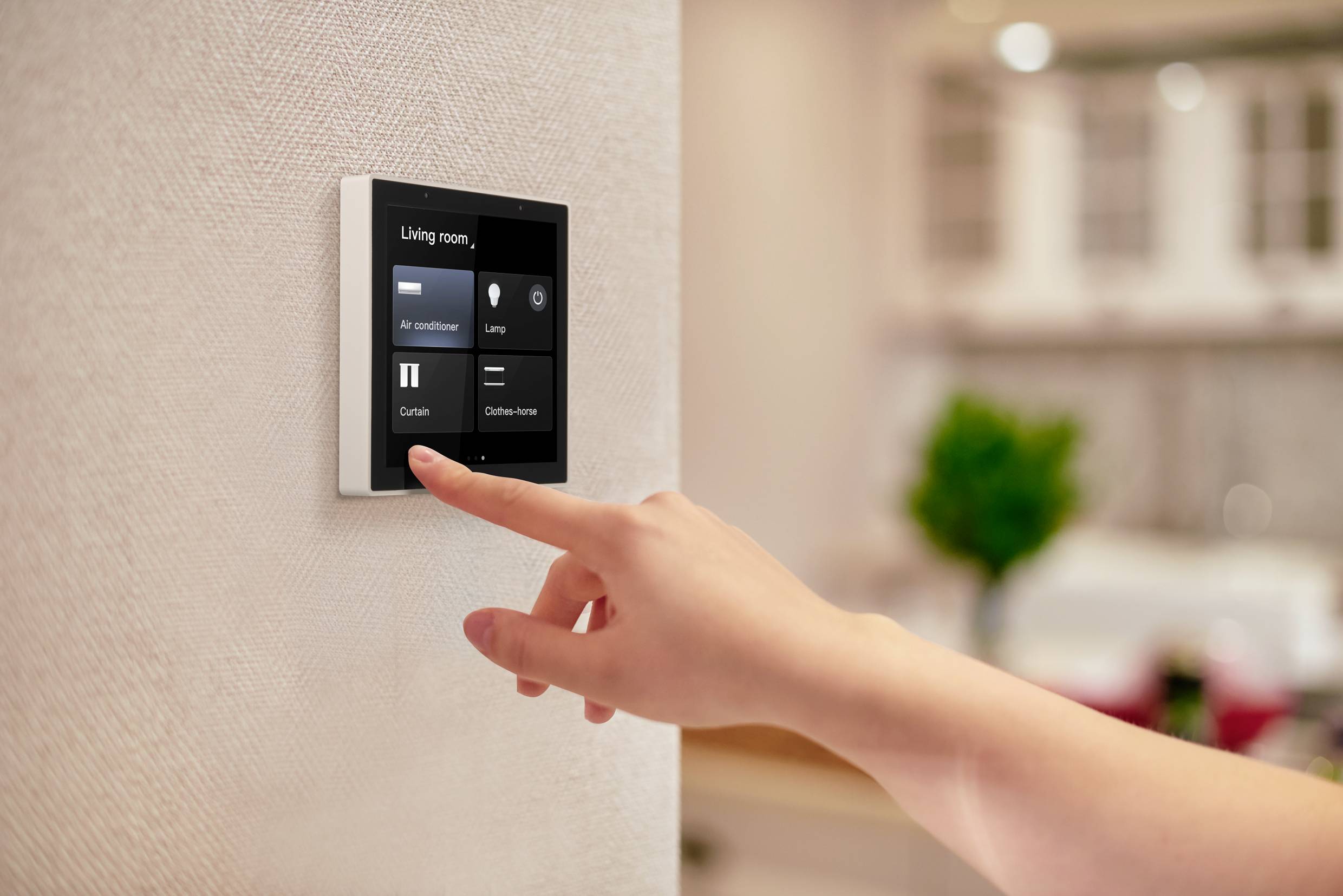 Eine Hand bedient ein digitales Smart-Home-Display an der Wand, um Geräte im Wohnzimmer zu steuern, wie Klimaanlage und Vorhänge.