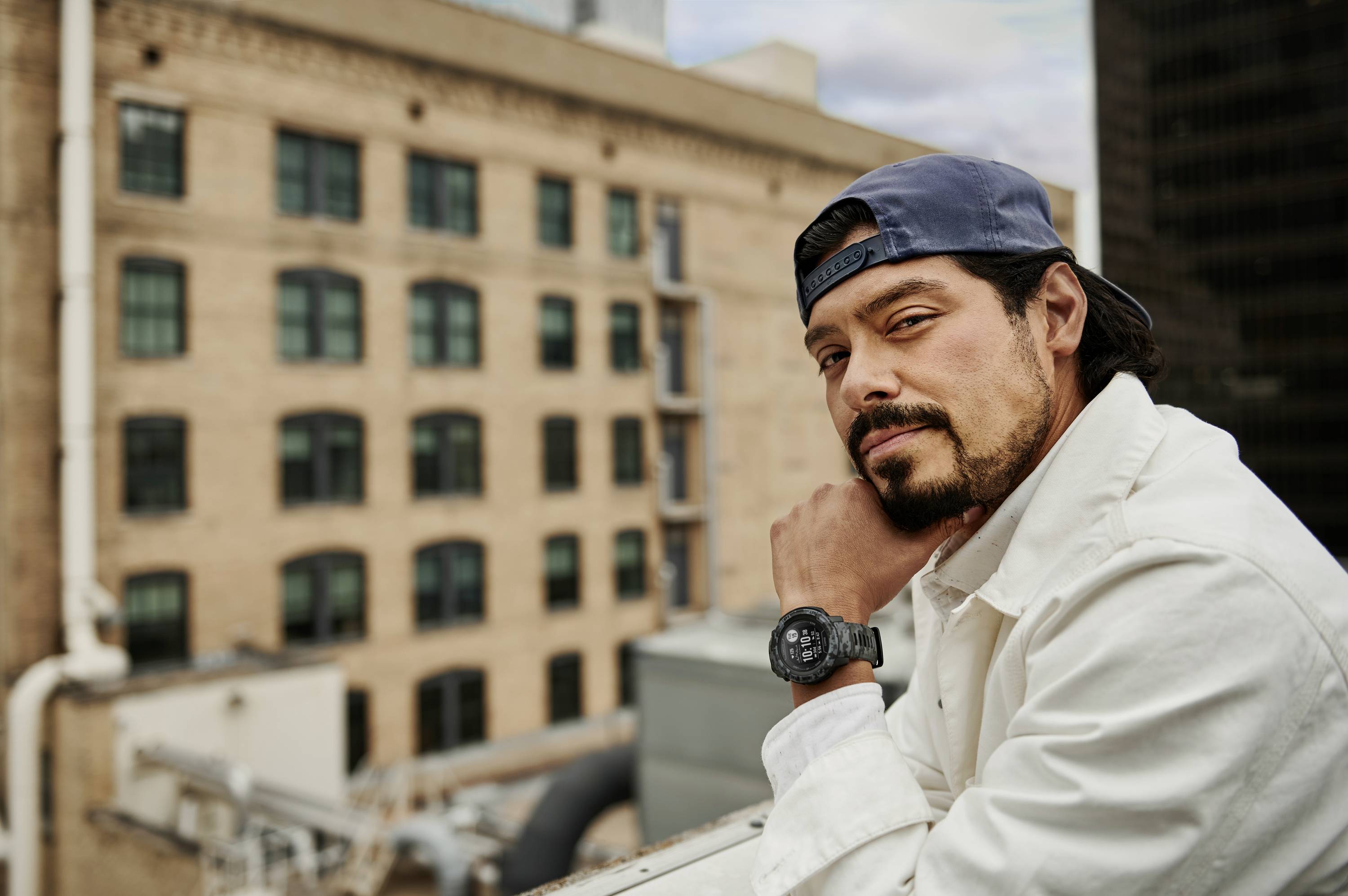 Ein Mann in einer weißen Jacke und blauer Kappe lehnt auf einem Geländer in einer städtischen Umgebung mit Backsteingebäuden im Hintergrund.