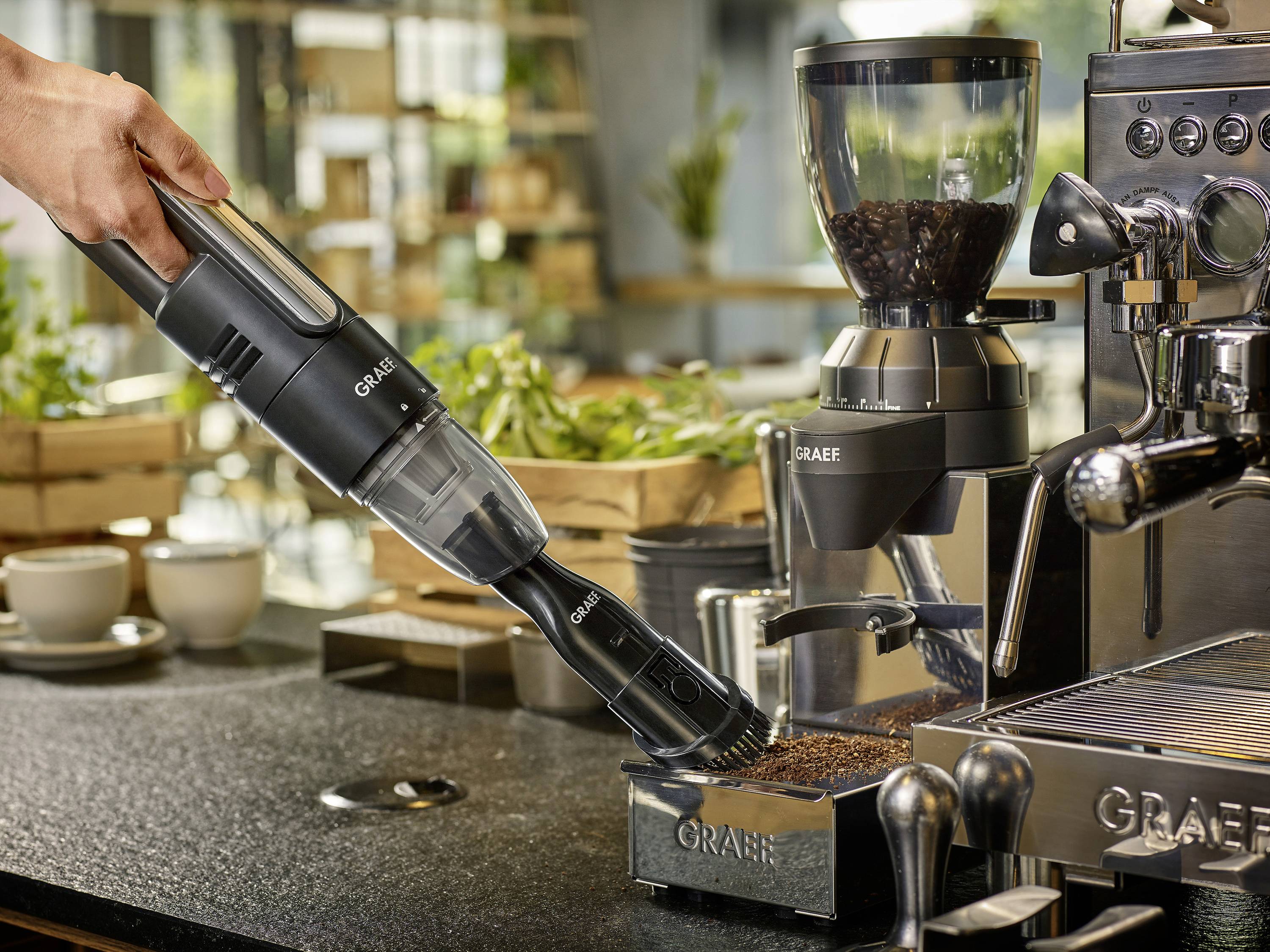 Eine Handstaubsauger saugt Kaffeemehl neben einer Kaffeemaschine auf. Im Hintergrund sind Pflanzen und Geschirr zu sehen.