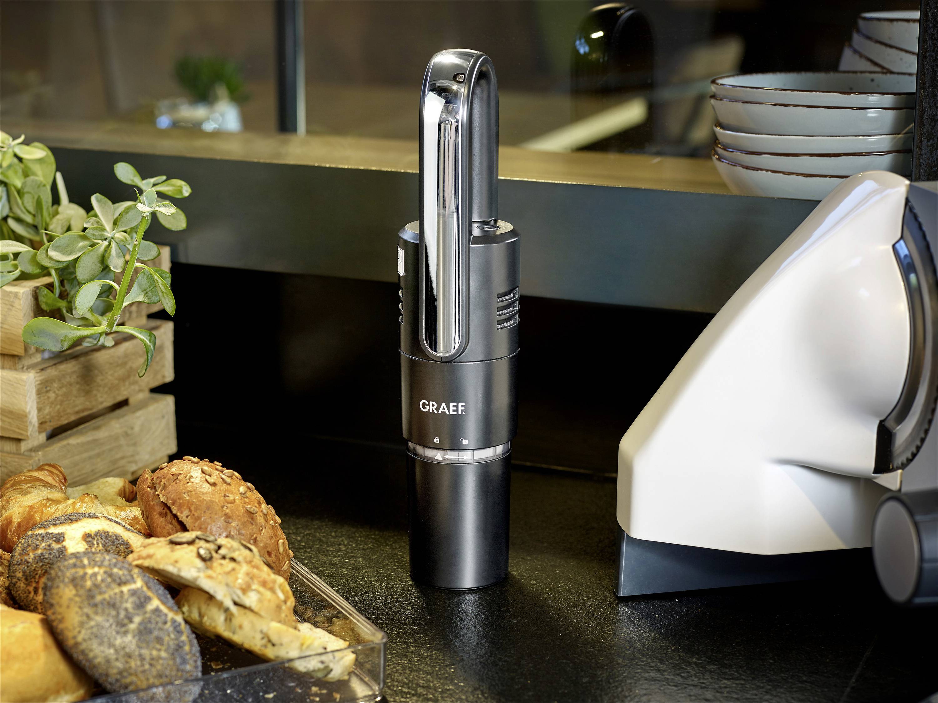 Ein elektrisches Graef-Messer steht auf einer Küchenarbeitsplatte neben einem Brotkorb und einem Brotschneider, umgeben von Geschirr.