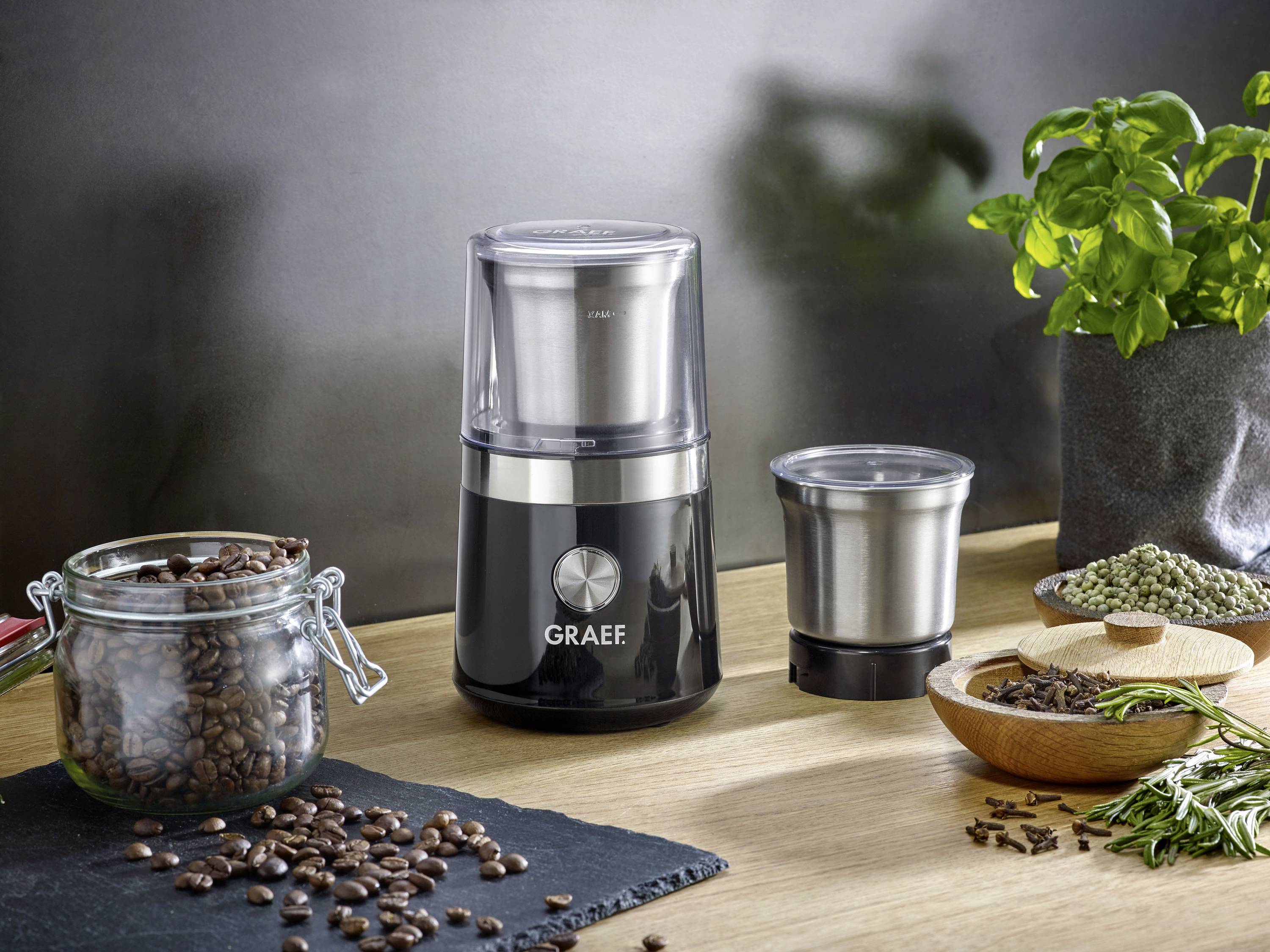 Elektrische Kaffeemühle auf einem Tisch mit Kaffeebohnen, Gewürzen und einer Pflanze daneben; modern und praktisch gestaltet.