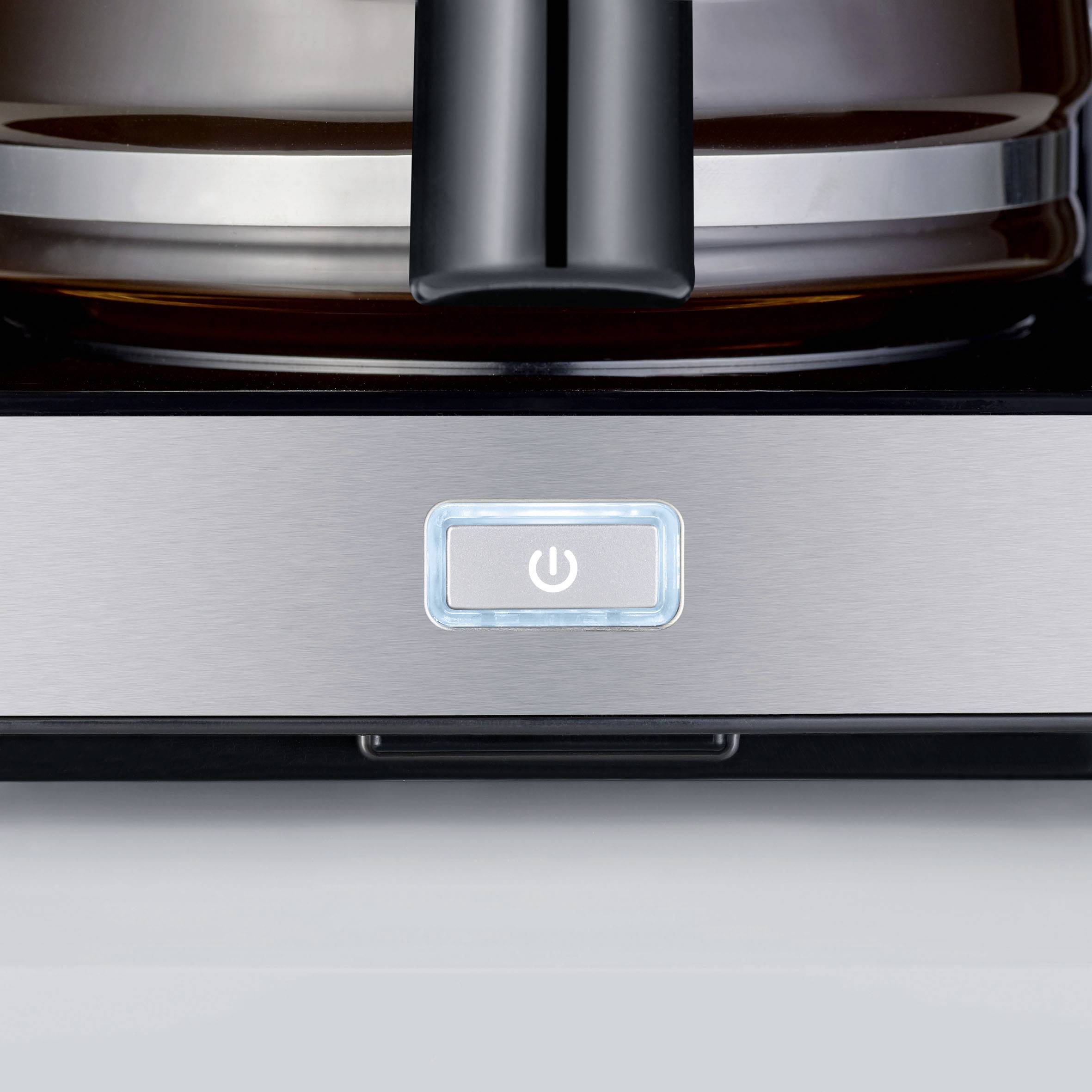 Kaffeemaschine mit einem beleuchteten Ein-/Ausschalter auf silbernem Gehäuse; Ausschnitt des Geräts zeigt Kanne im Hintergrund.