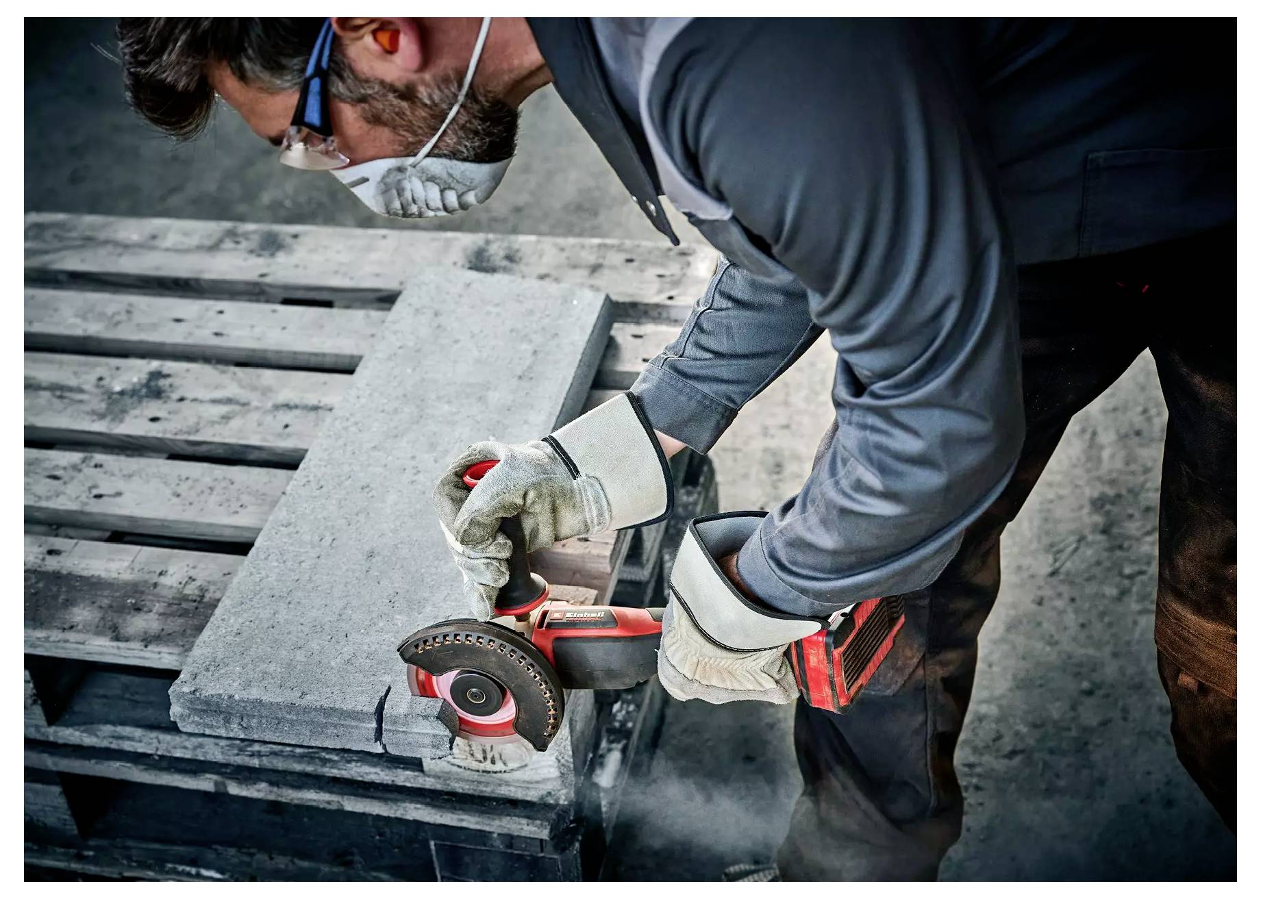 Eine Person in Schutzausrüstung schleift einen Betonblock mit einem Elektrowerkzeug auf einer Holzpalette und veranschaulicht damit die Werkzeuge, die in der Bauarbeit verwendet werden.